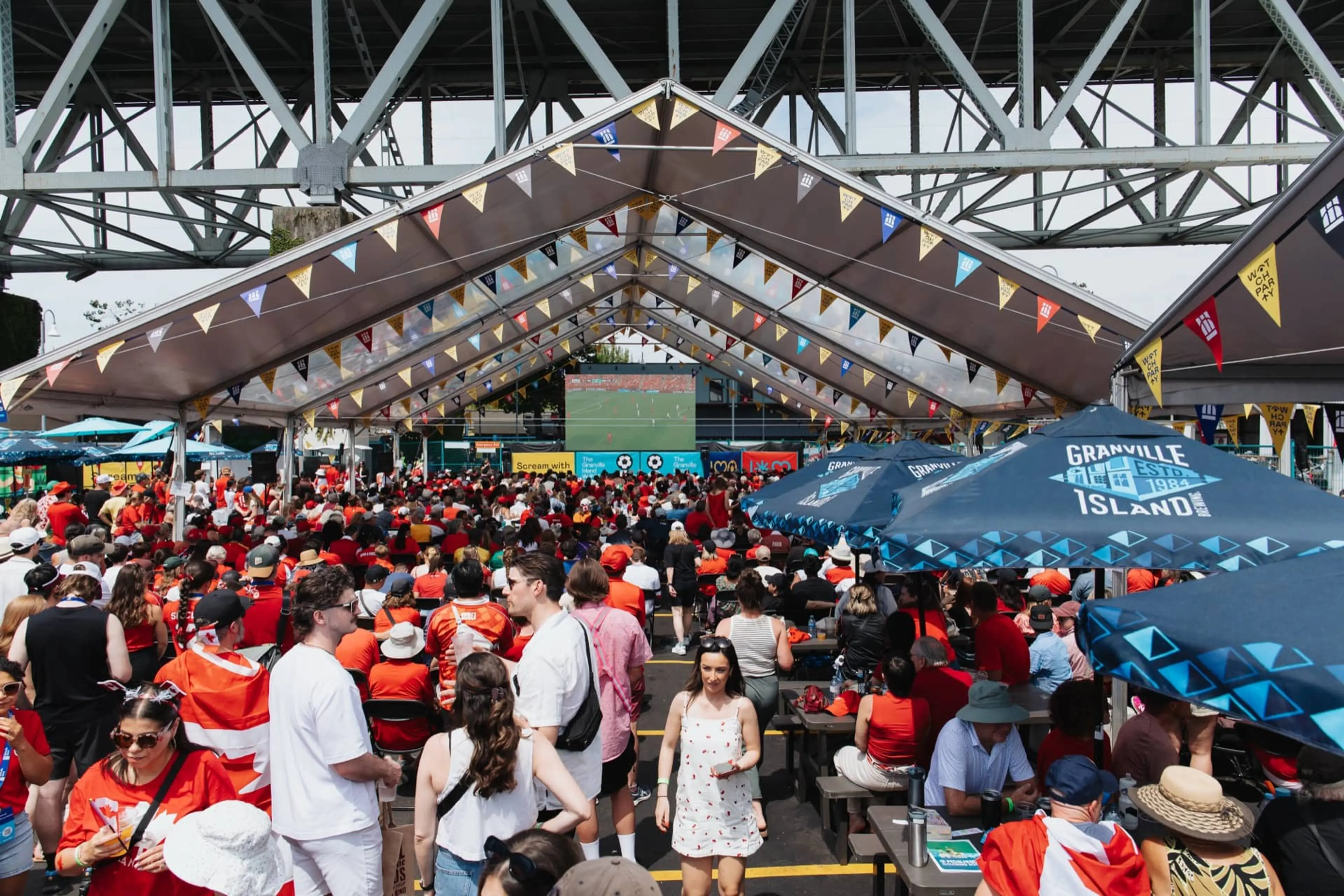 Crowd of people under a large tent with colorful flags watching a sports game on a big screen under a bridge.