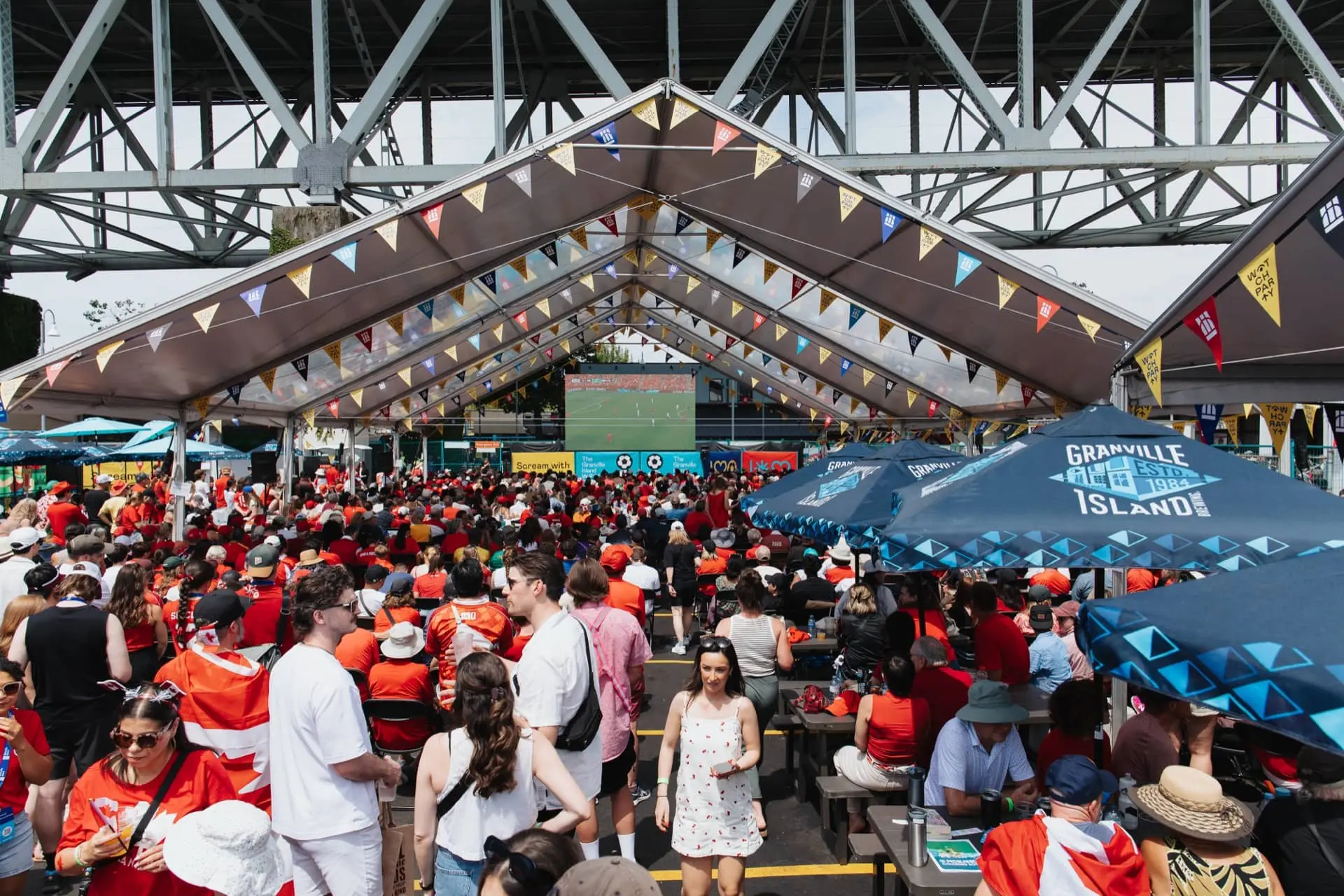 Crowd of people under a large tent with colorful flags watching a sports game on a big screen under a bridge.