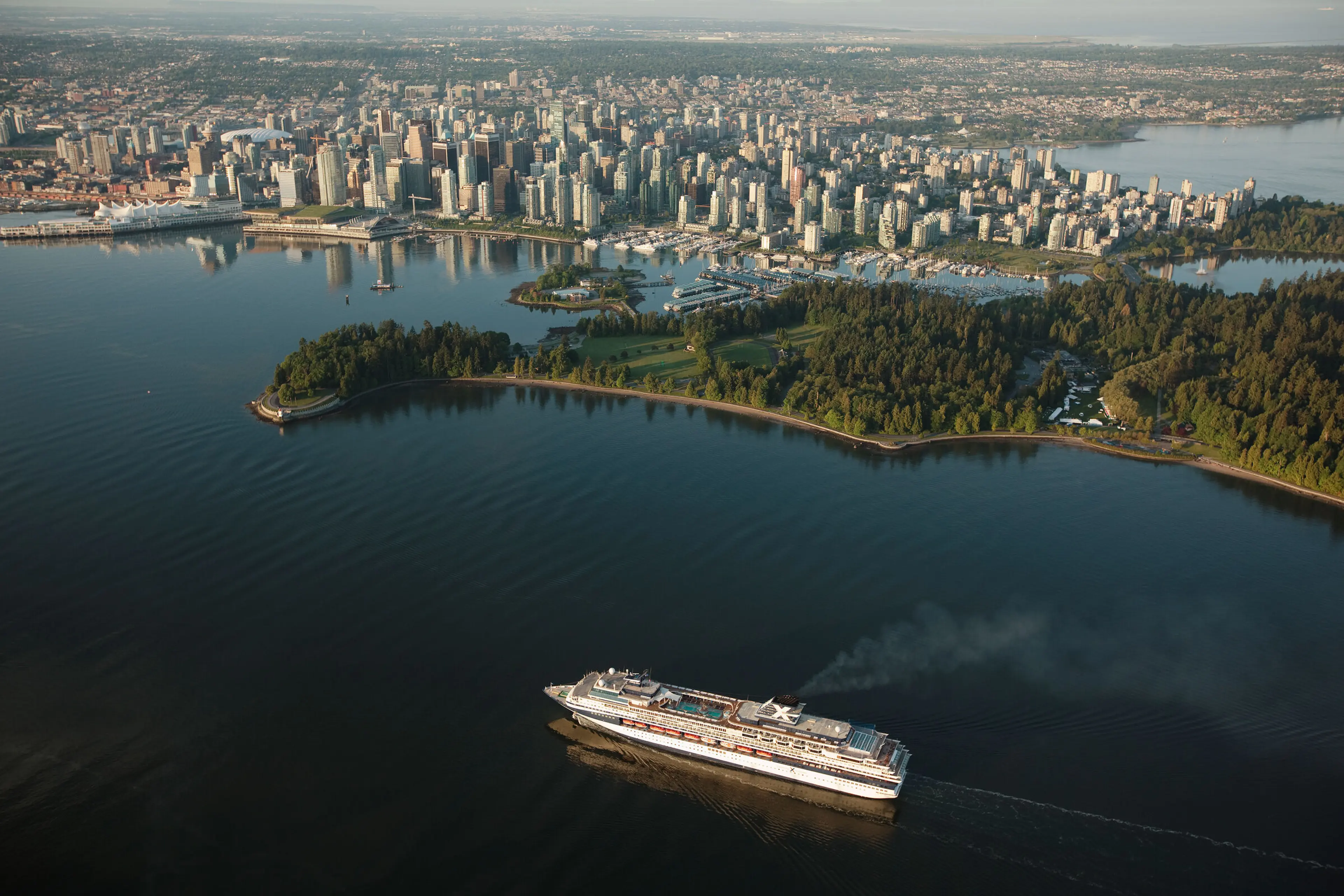 A large cruise ship sailing near a forested peninsula with a city skyline in the background across the water.