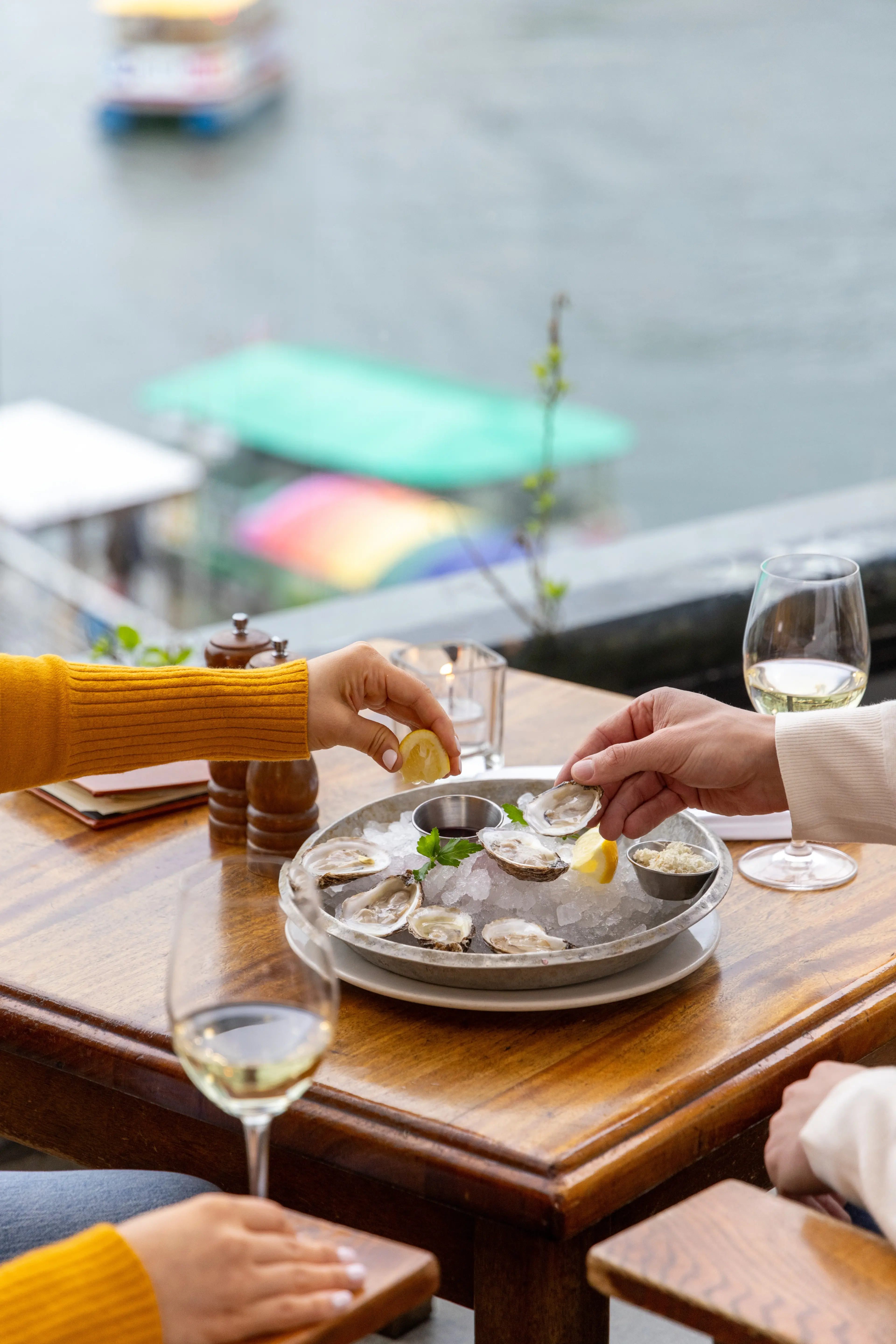 Two people at a wooden table by the water eating oysters on ice with lemon wedges and white wine glasses.
