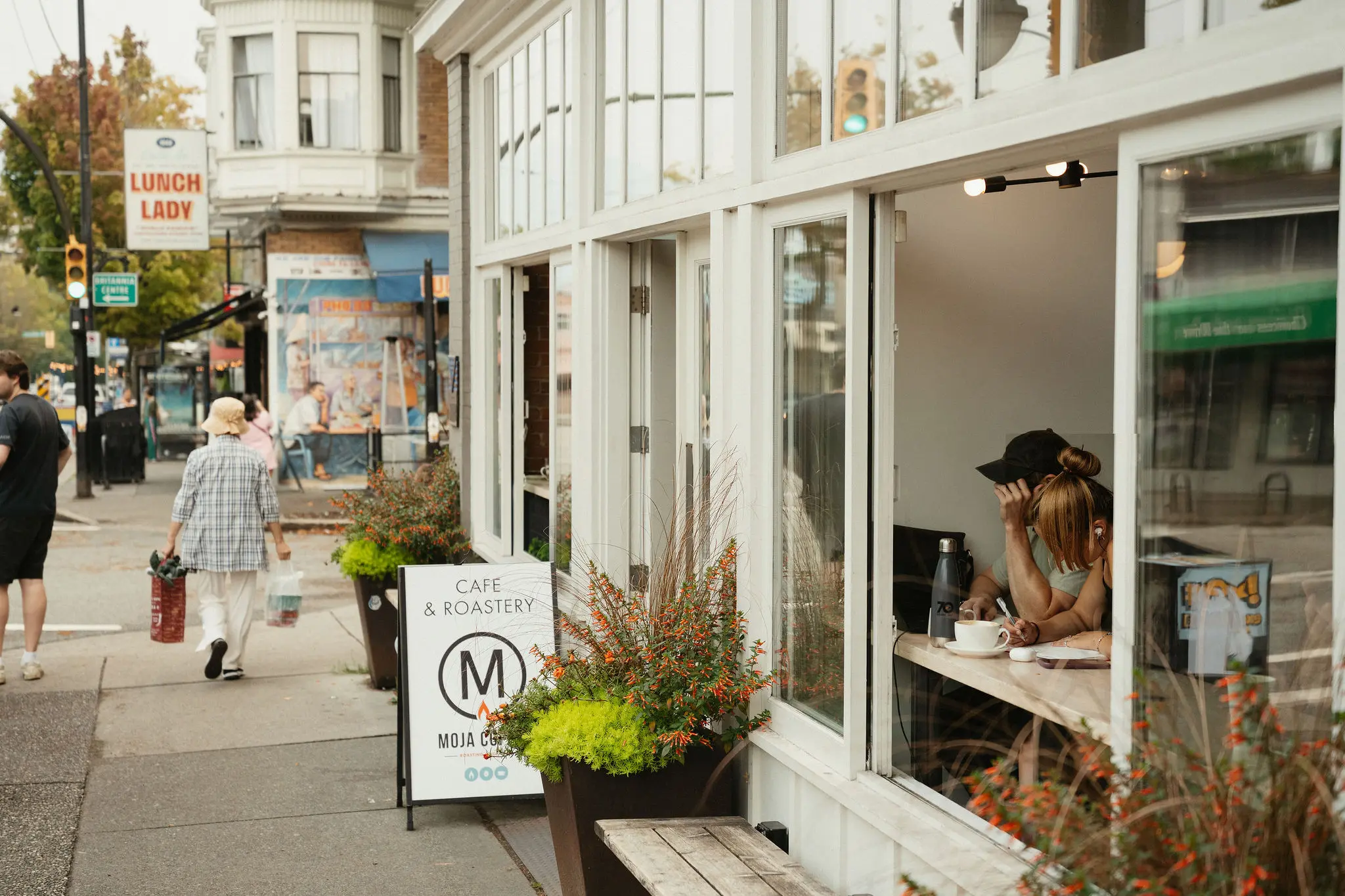 Two people sitting inside a cafe near an open window, with a sign outside reading Cafe & Roastery Moja Coffee.