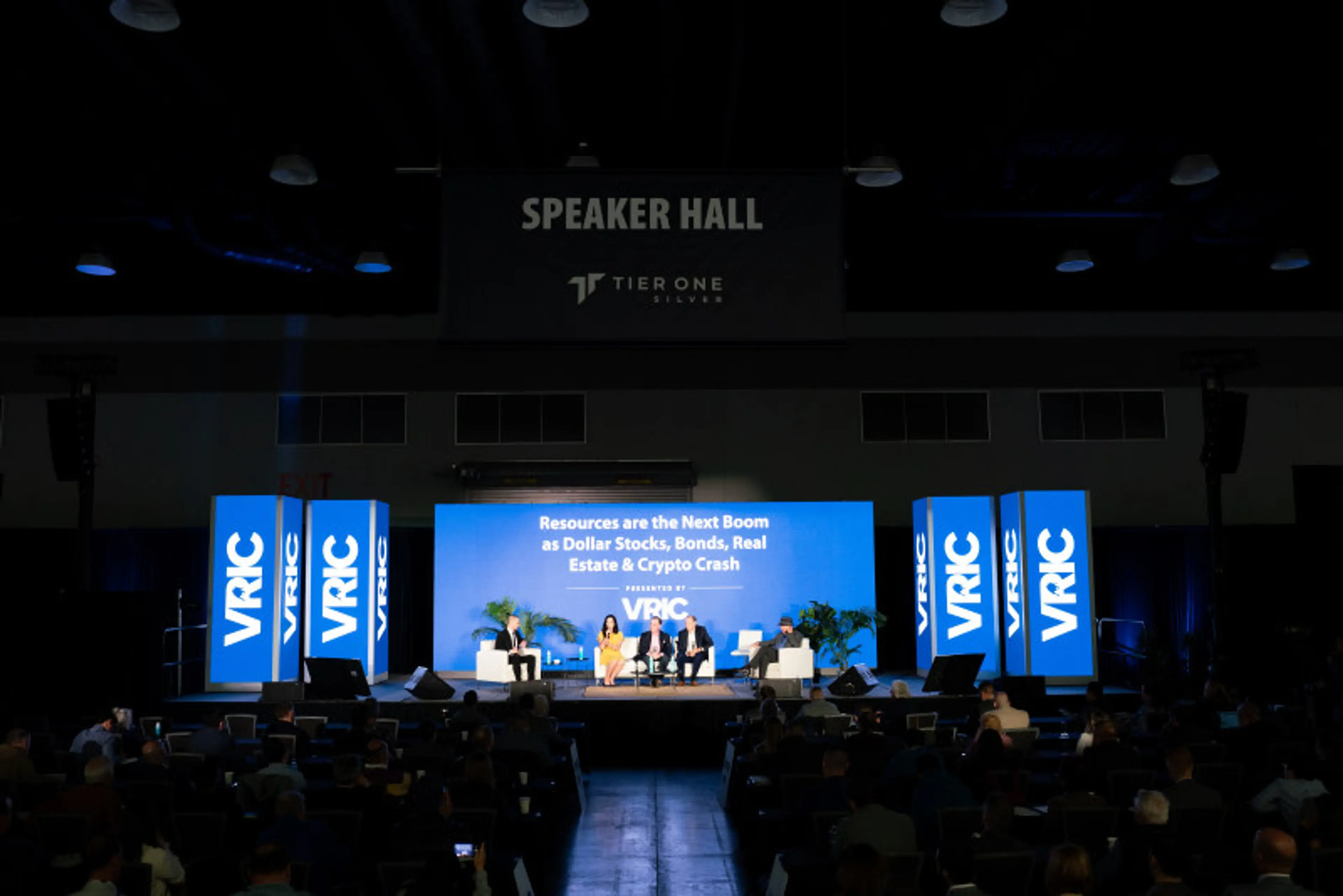 Panel discussion on stage with four seated speakers and large blue screens displaying VRIC and a topic about resources and market crash.