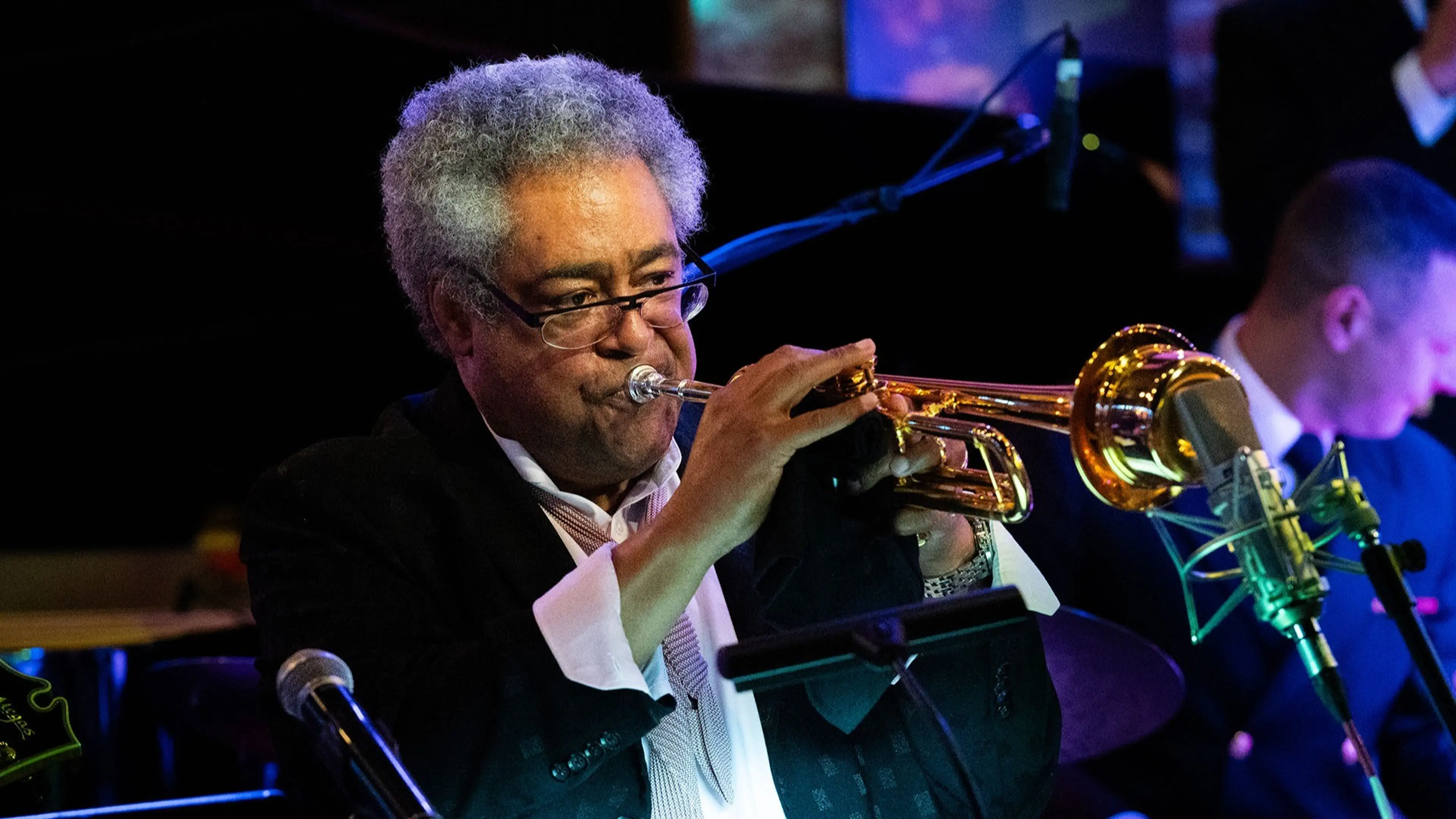 A person with gray curly hair and glasses playing a trumpet on stage with microphones nearby.