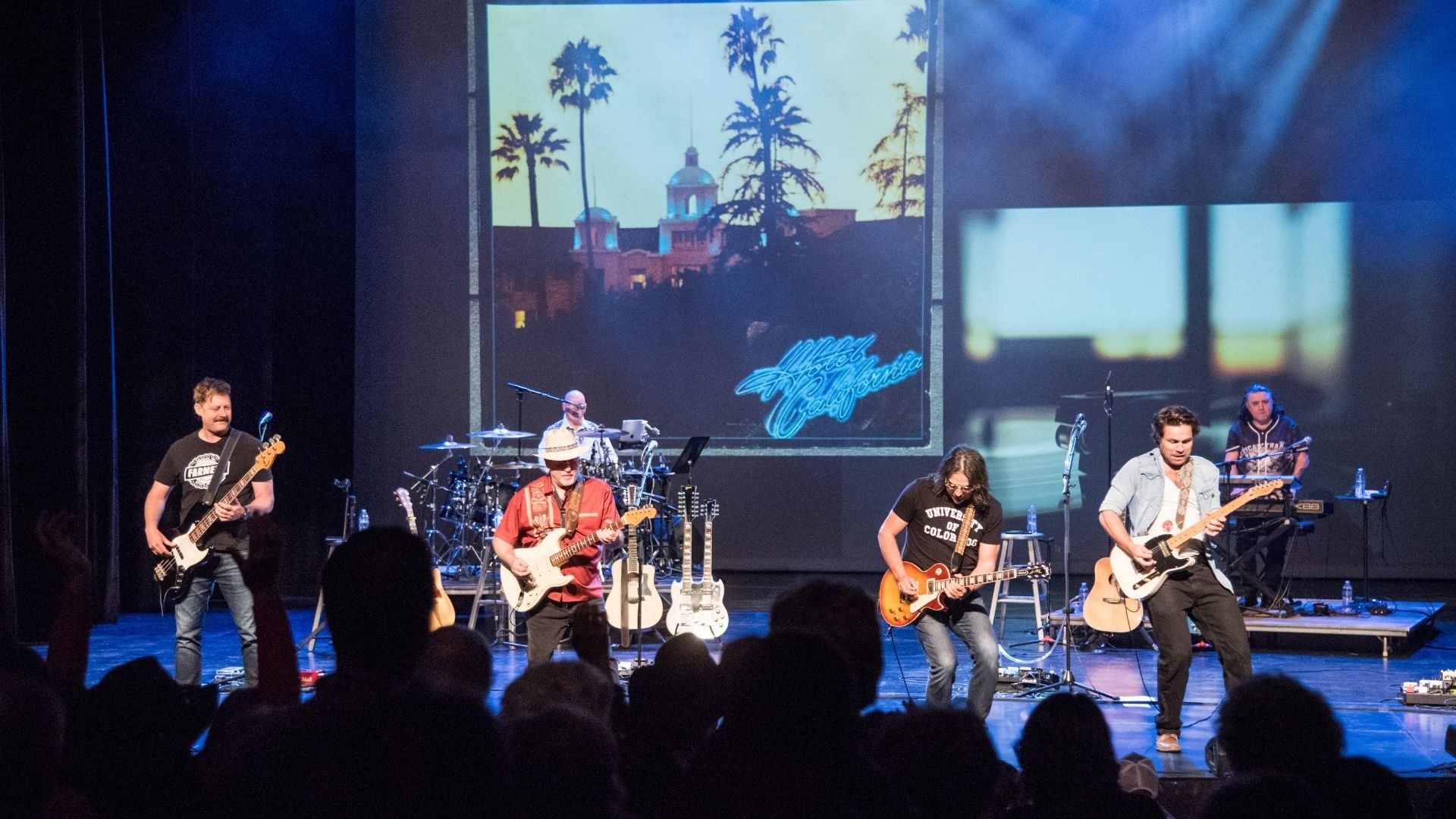 Five musicians performing on stage with guitars, drums, and keyboard, with a large screen showing a sunset and palm trees behind them.