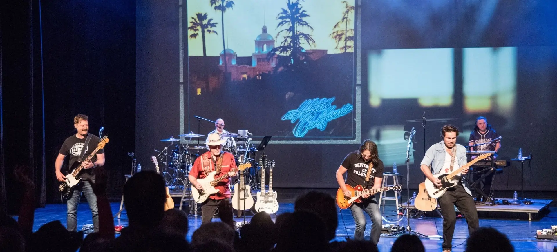 Five musicians performing on stage with guitars, drums, and keyboard, with a large screen showing a sunset and palm trees behind them.