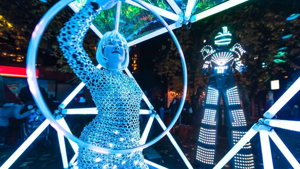 Person in a shiny silver outfit and futuristic glasses holding a glowing hoop, with a tall figure in a light-up suit in the background at night.