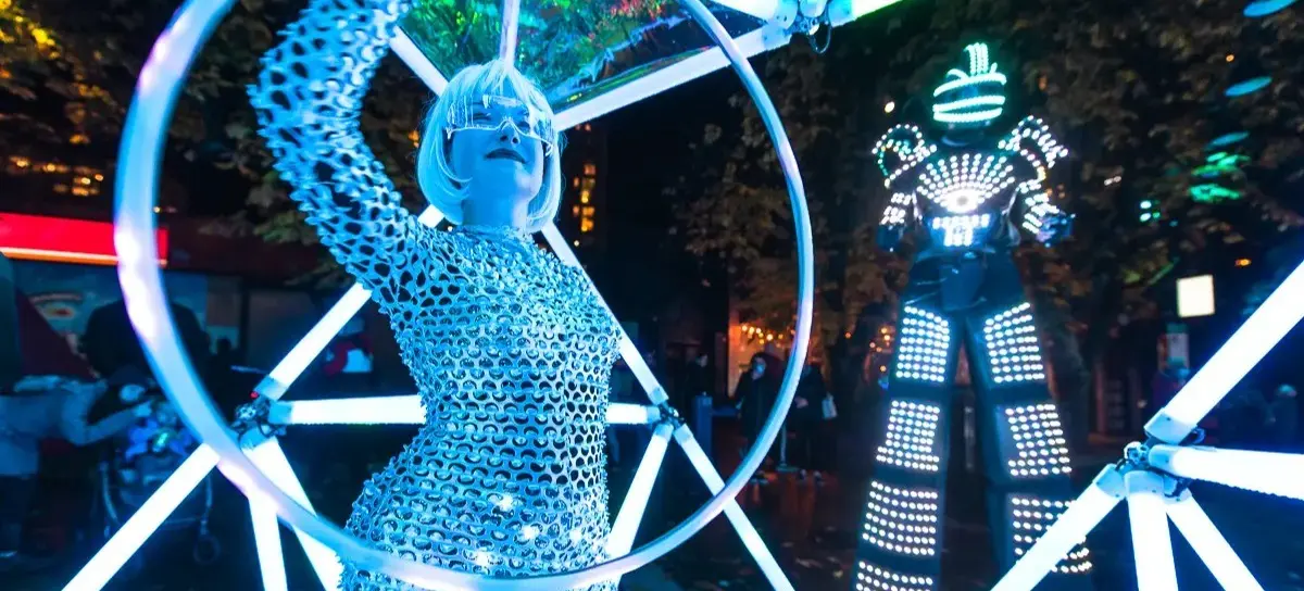 Person in a shiny silver outfit and futuristic glasses holding a glowing hoop, with a tall figure in a light-up suit in the background at night.