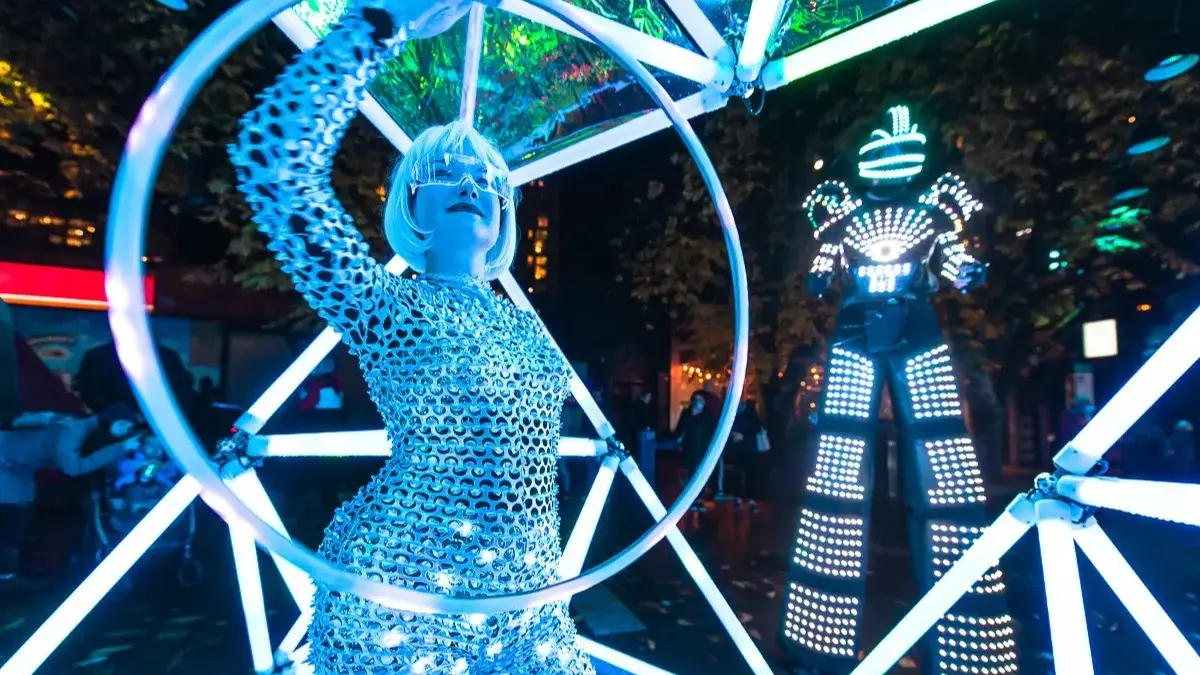 Person in a shiny silver outfit and futuristic glasses holding a glowing hoop, with a tall figure in a light-up suit in the background at night.