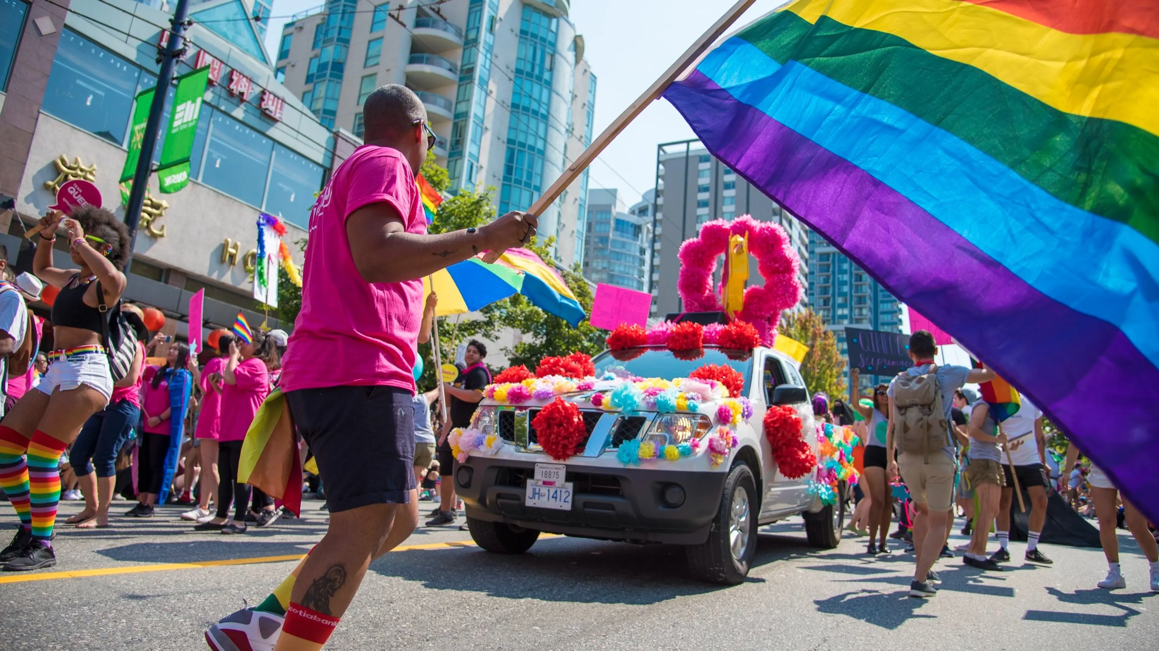Person in pink shirt holding a large rainbow flag near a decorated vehicle at a colorful pride parade with people wearing rainbow accessories.