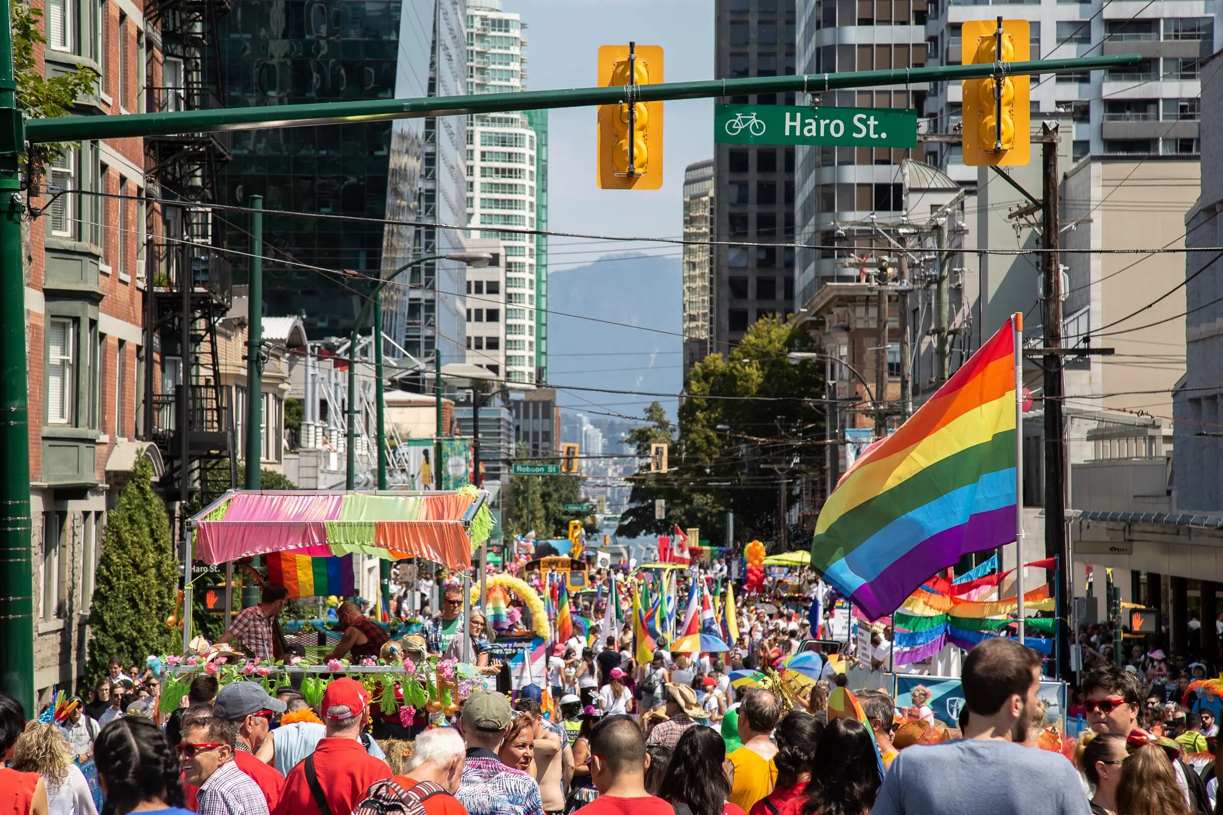 Crowded street parade with rainbow flags and colorful decorations under a Haro St. sign in an urban area.