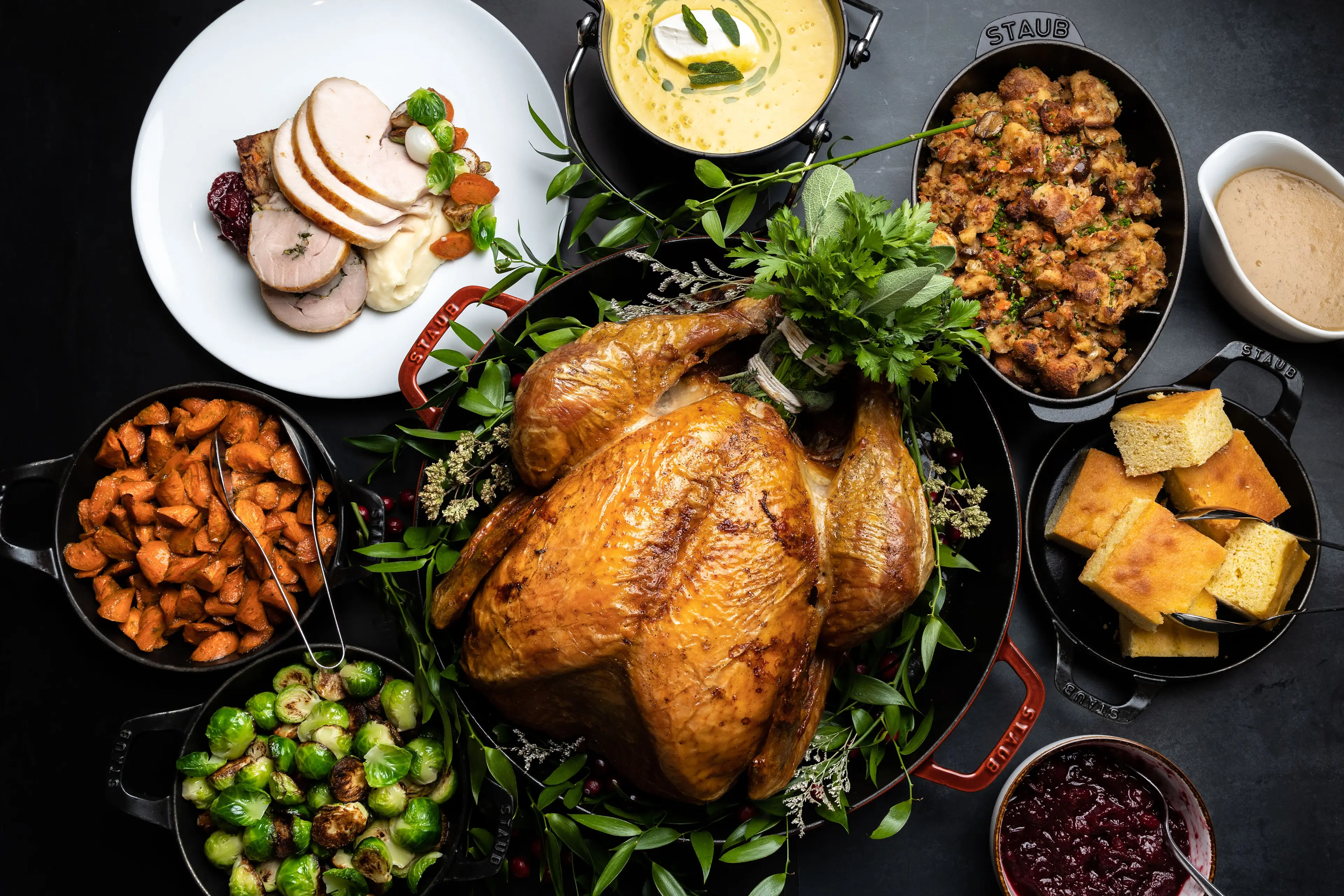 Roasted whole turkey garnished with herbs surrounded by dishes of sliced turkey, mashed potatoes, stuffing, cornbread, roasted carrots, Brussels sprouts, and cranberry sauce.
