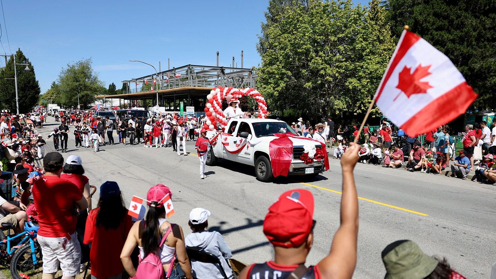 Crowd watching a parade with a white truck decorated with red and white balloons and Canadian flags on a sunny day.