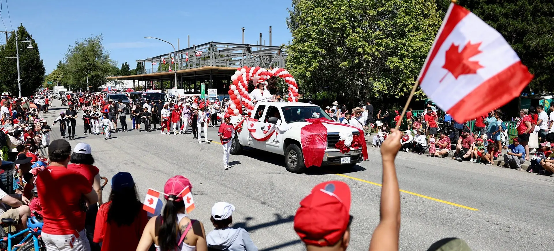 Crowd watching a parade with a white truck decorated with red and white balloons and Canadian flags on a sunny day.