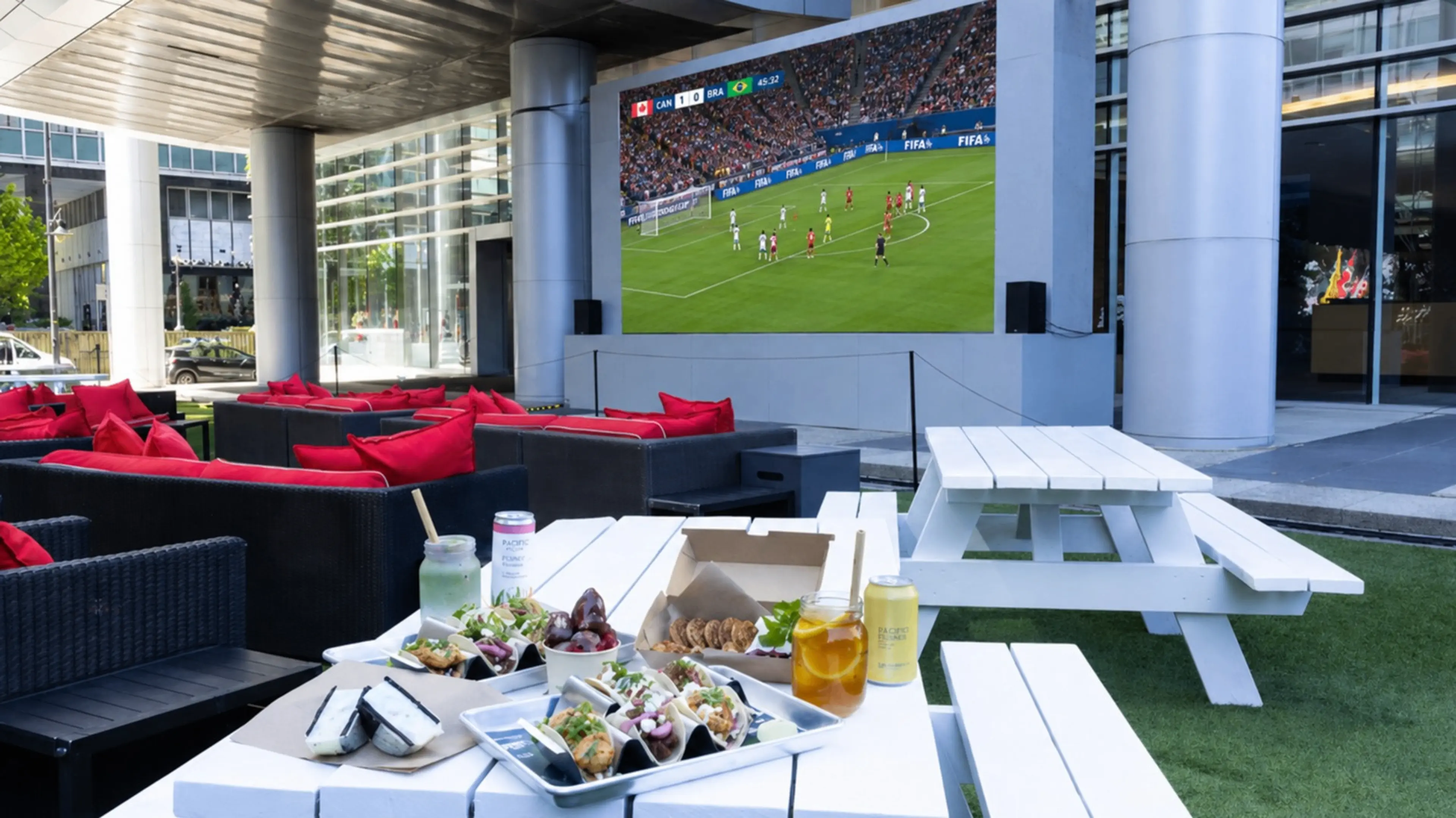 Outdoor seating area with white picnic tables and black couches with red pillows, food and drinks on a table, and a large screen showing a soccer match.