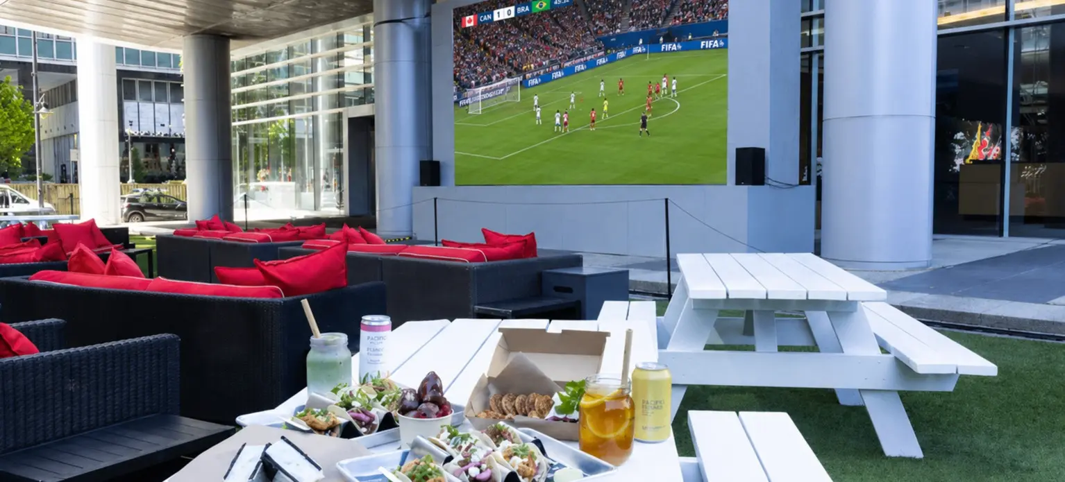 Outdoor seating area with white picnic tables and black couches with red pillows, food and drinks on a table, and a large screen showing a soccer match.