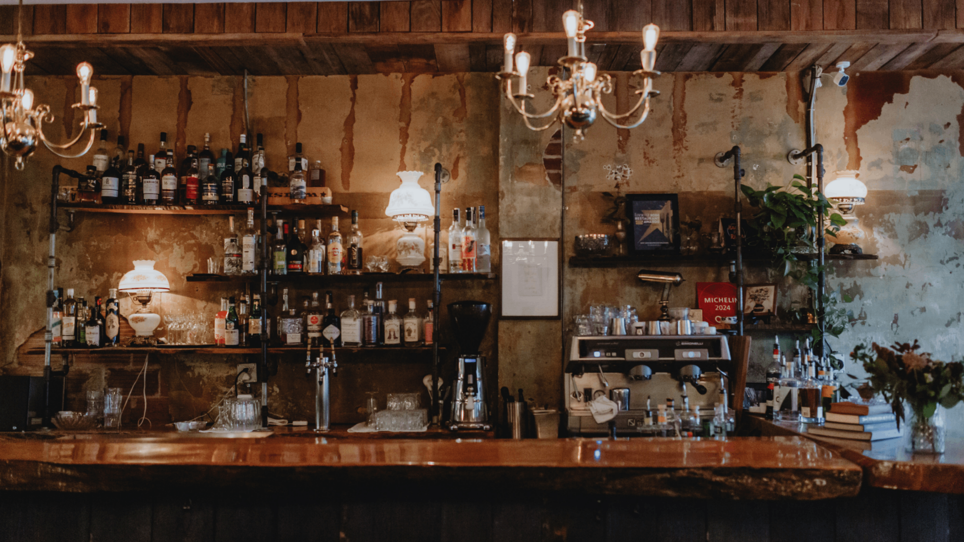 Rustic bar counter with shelves of liquor bottles, glassware, coffee machine, and warm hanging lights.