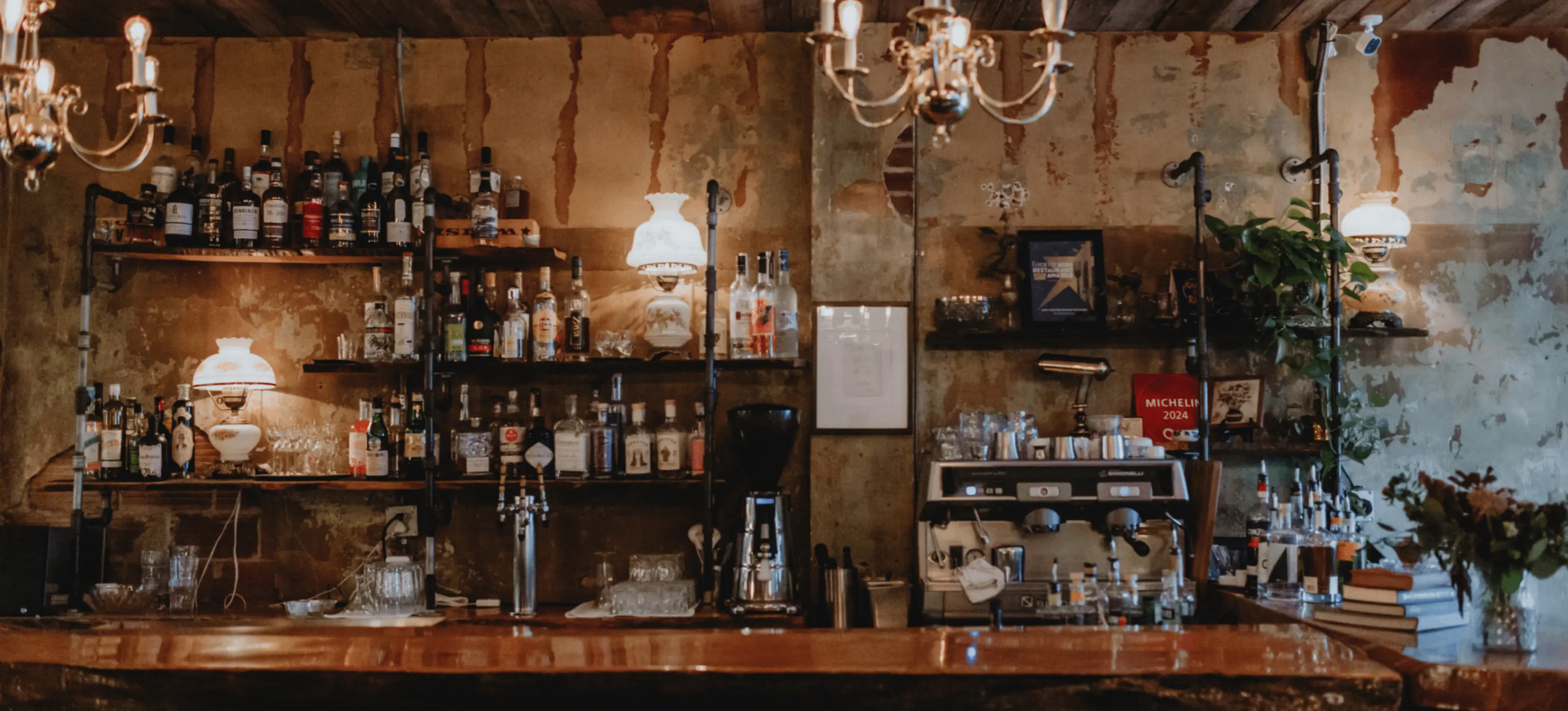 Rustic bar counter with shelves of liquor bottles, glassware, coffee machine, and warm hanging lights.