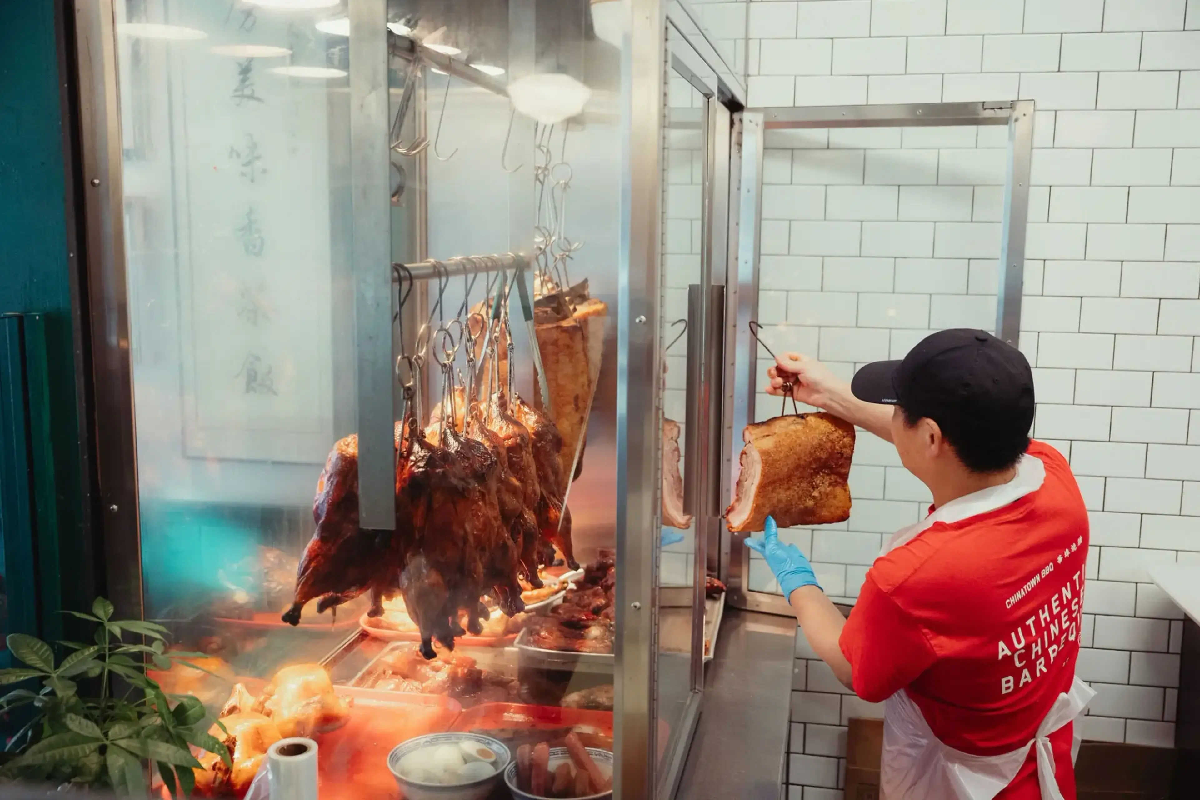 Person in red shirt and black cap holding roasted meat inside a glass display with hanging roasted ducks and pork.