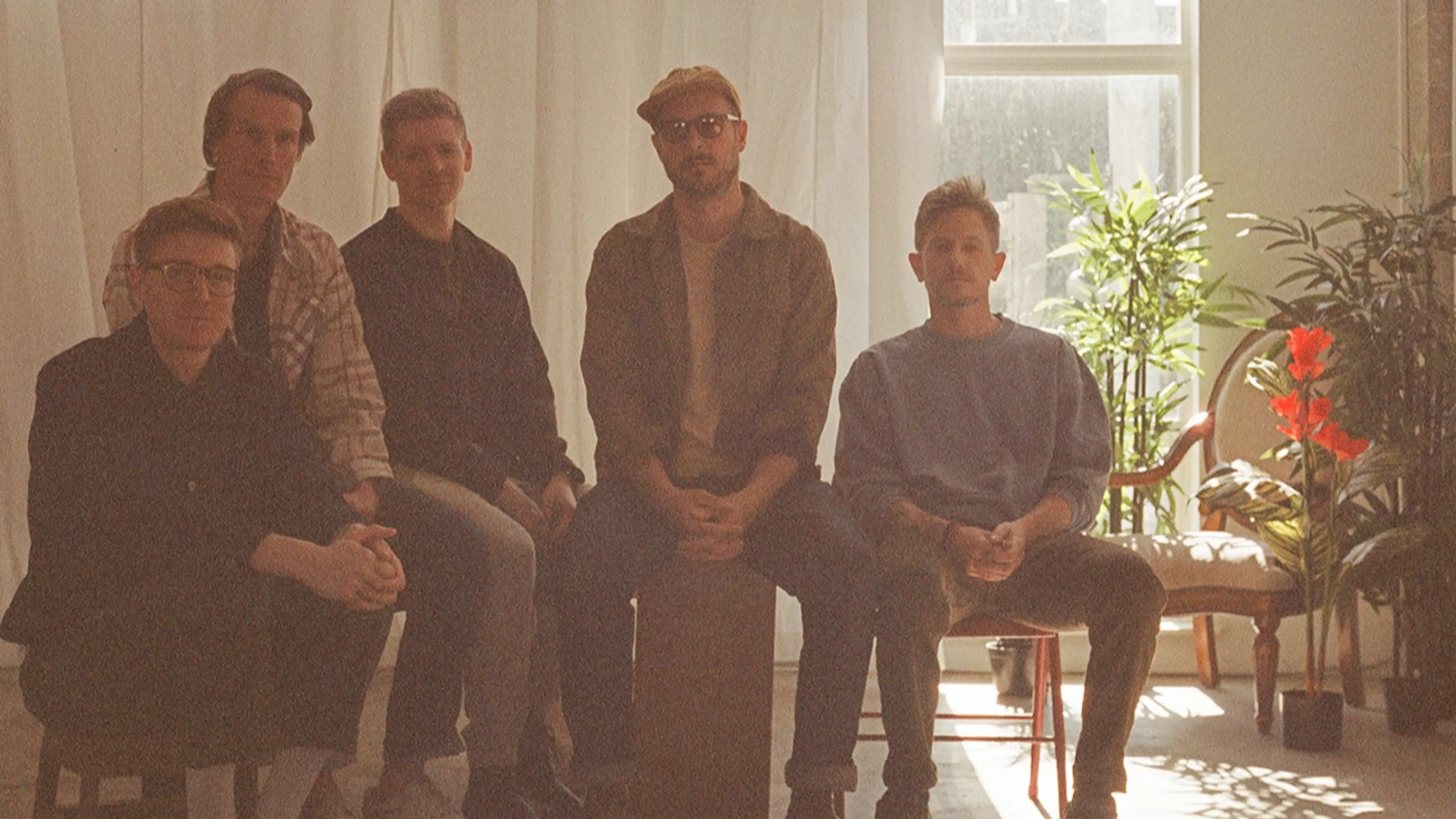 Five people sitting indoors with plants and sunlight in the background, some wearing glasses and casual clothing.
