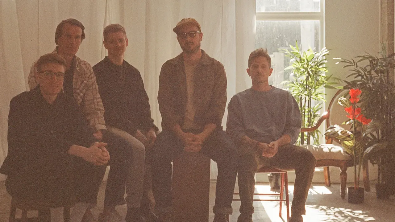 Five people sitting indoors with plants and sunlight in the background, some wearing glasses and casual clothing.