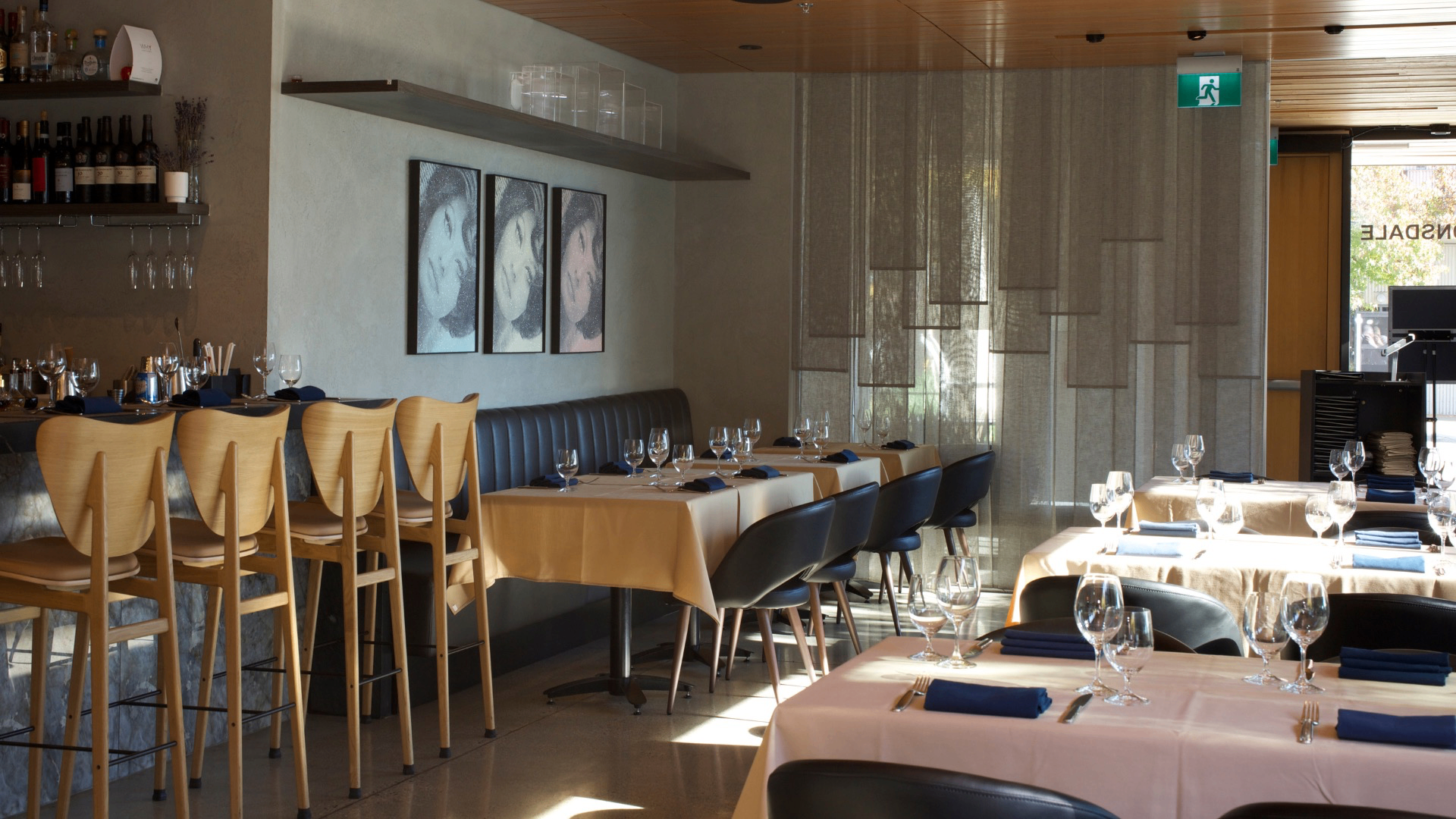 Empty restaurant interior with wooden bar stools, tables set with glasses and blue napkins, and three framed portraits on the wall.