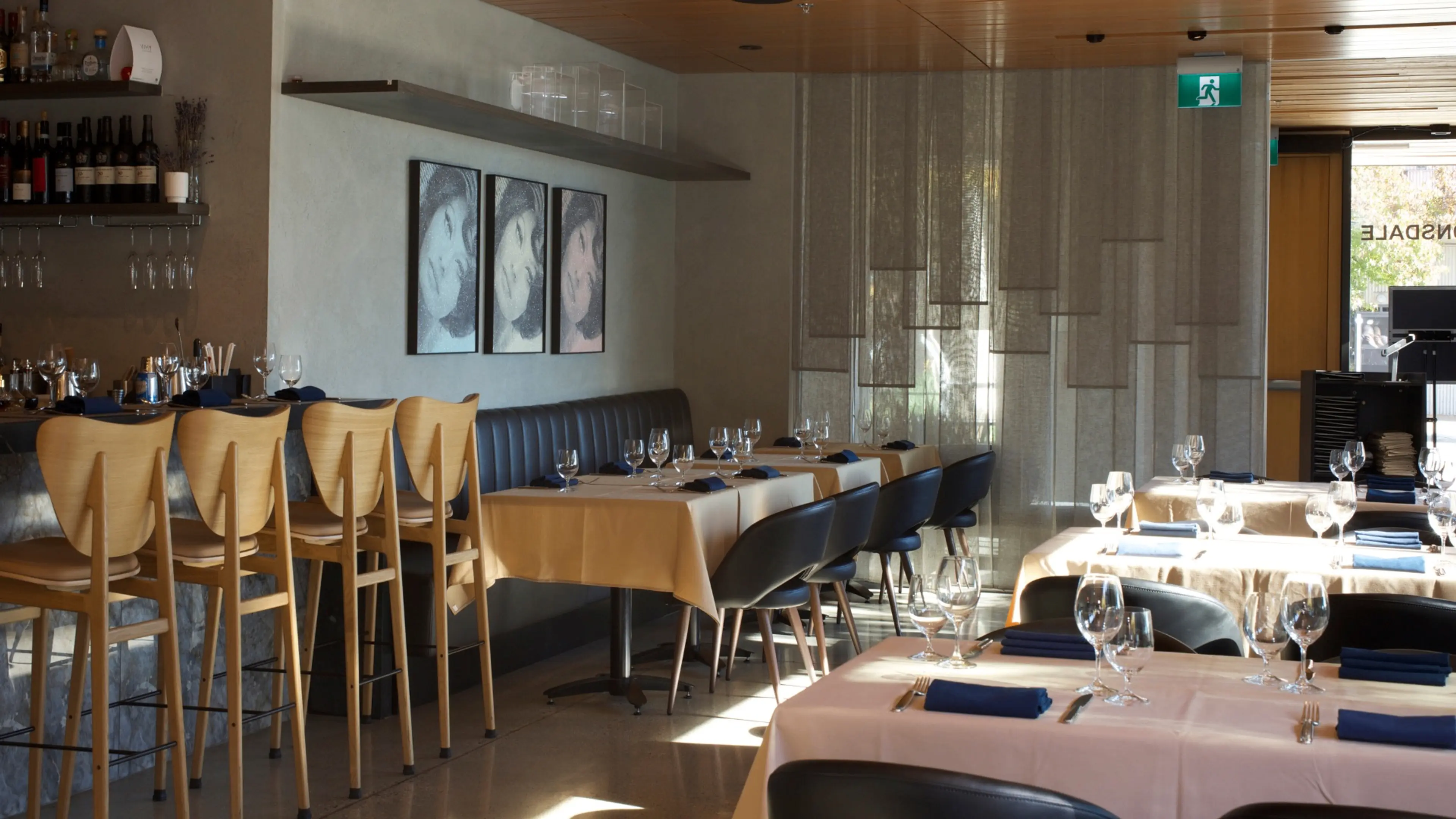 Empty restaurant interior with wooden bar stools, tables set with glasses and blue napkins, and three framed portraits on the wall.