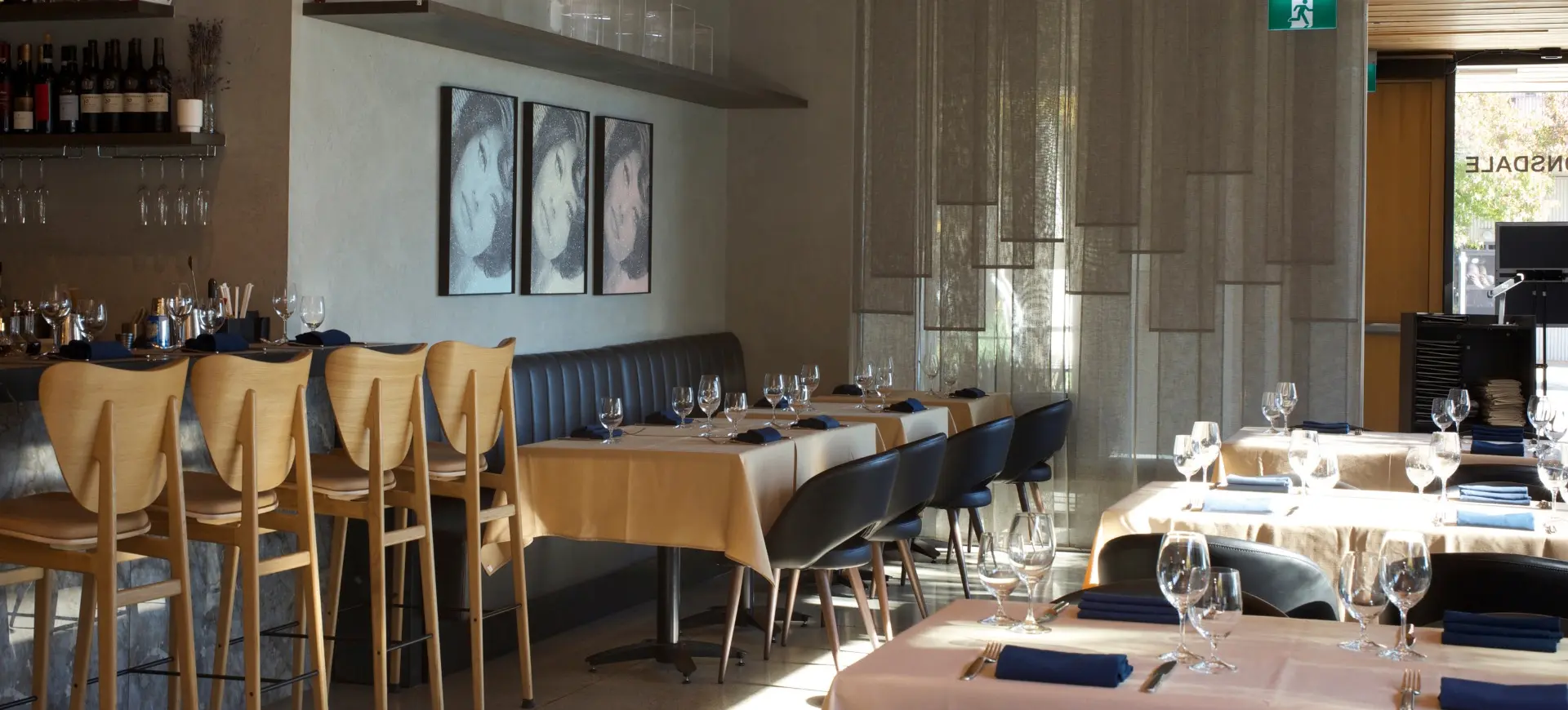 Empty restaurant interior with wooden bar stools, tables set with glasses and blue napkins, and three framed portraits on the wall.