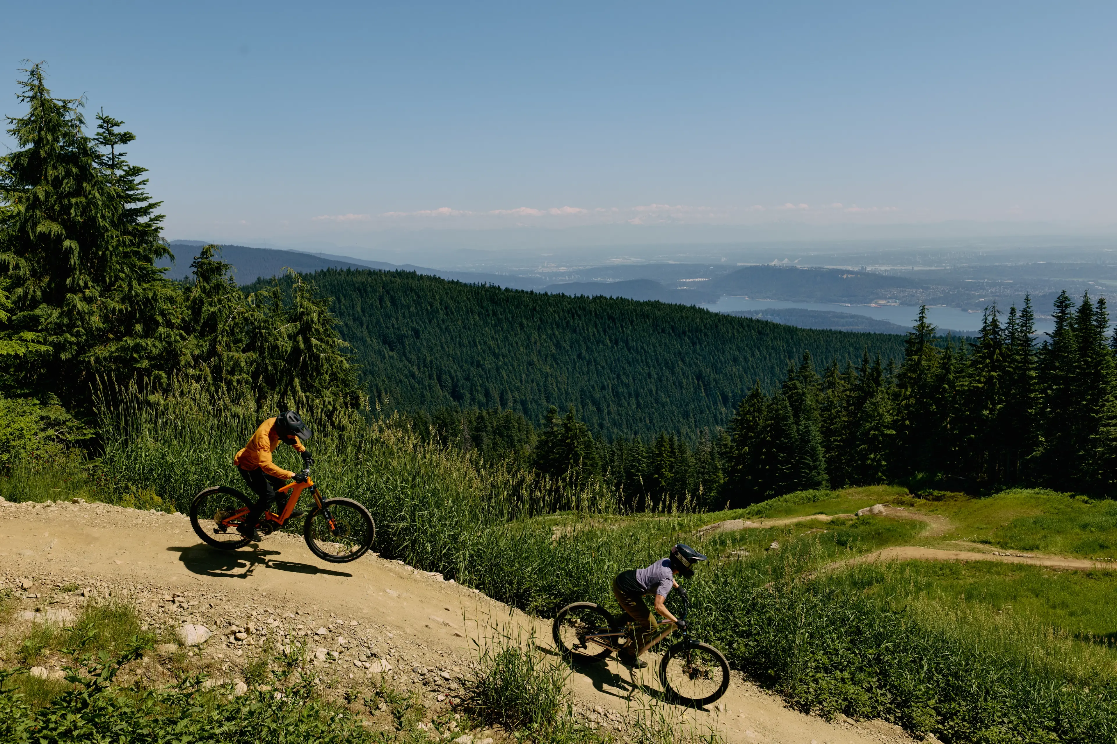 Two mountain bikers riding downhill on a dirt trail surrounded by green trees and hills under a clear blue sky.