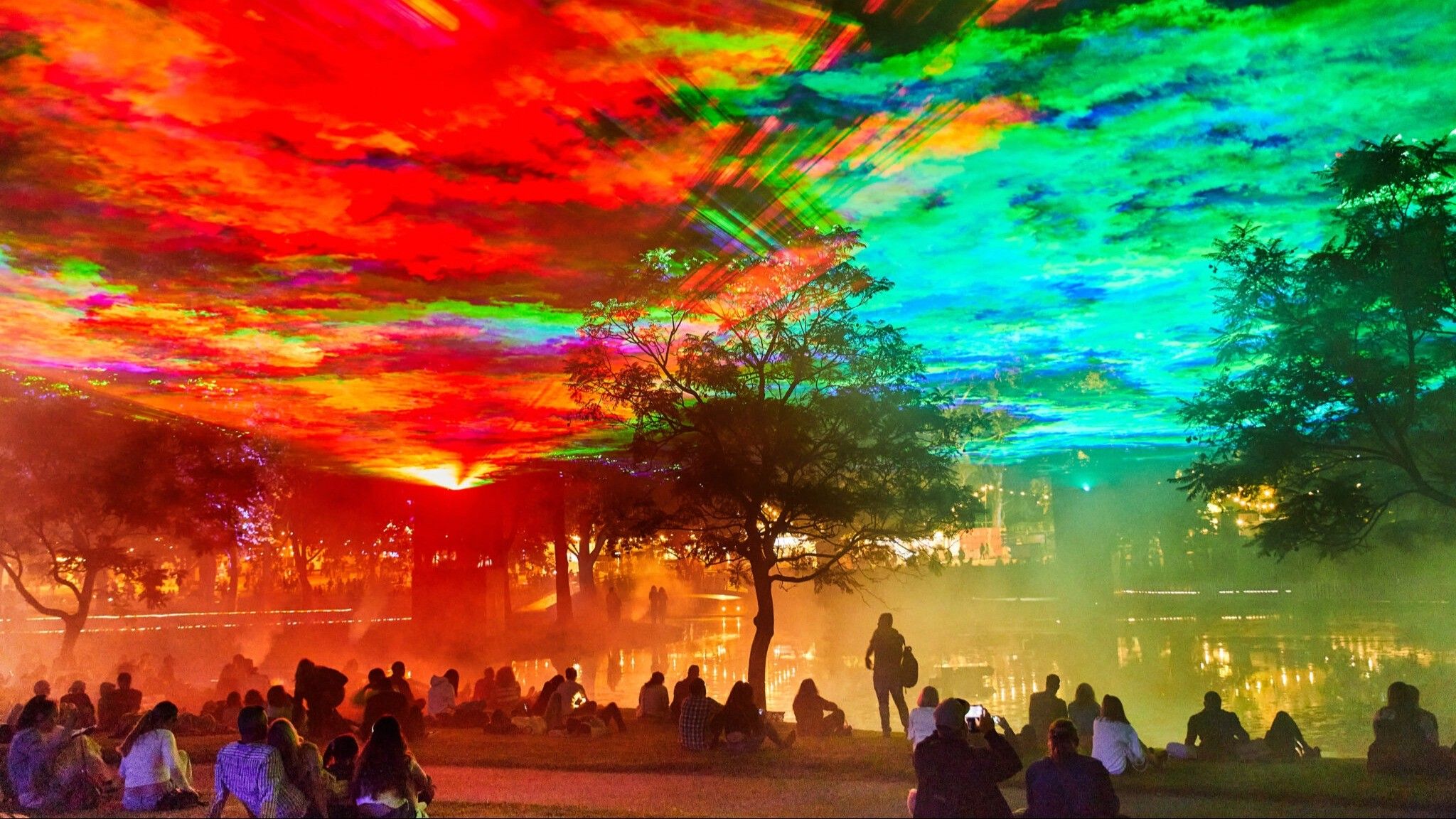 People sitting and standing near a lake under a colorful sky with red, green, and blue light effects and trees around.