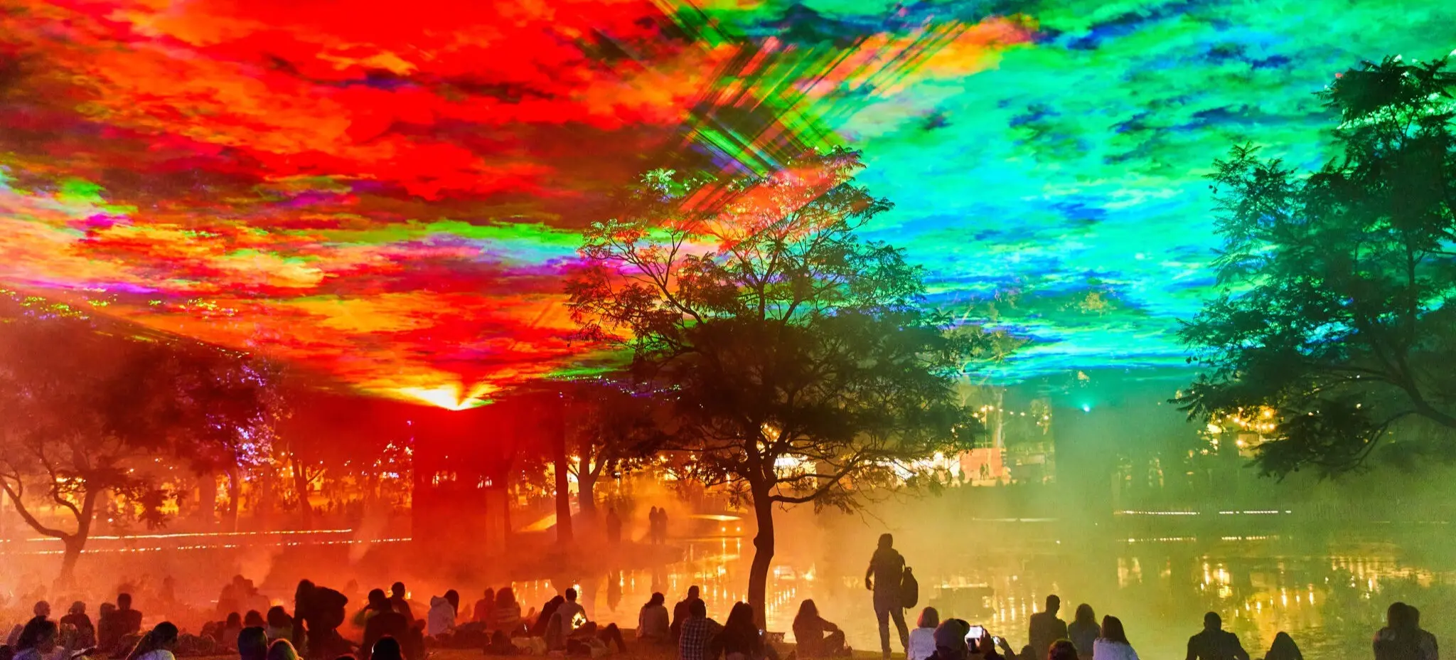 People sitting and standing near a lake under a colorful sky with red, green, and blue light effects and trees around.