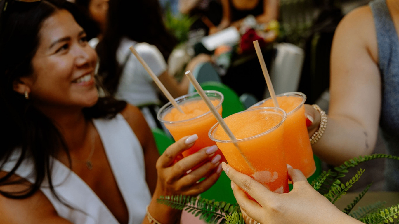 Three people holding and clinking plastic cups filled with orange slush drinks with straws.