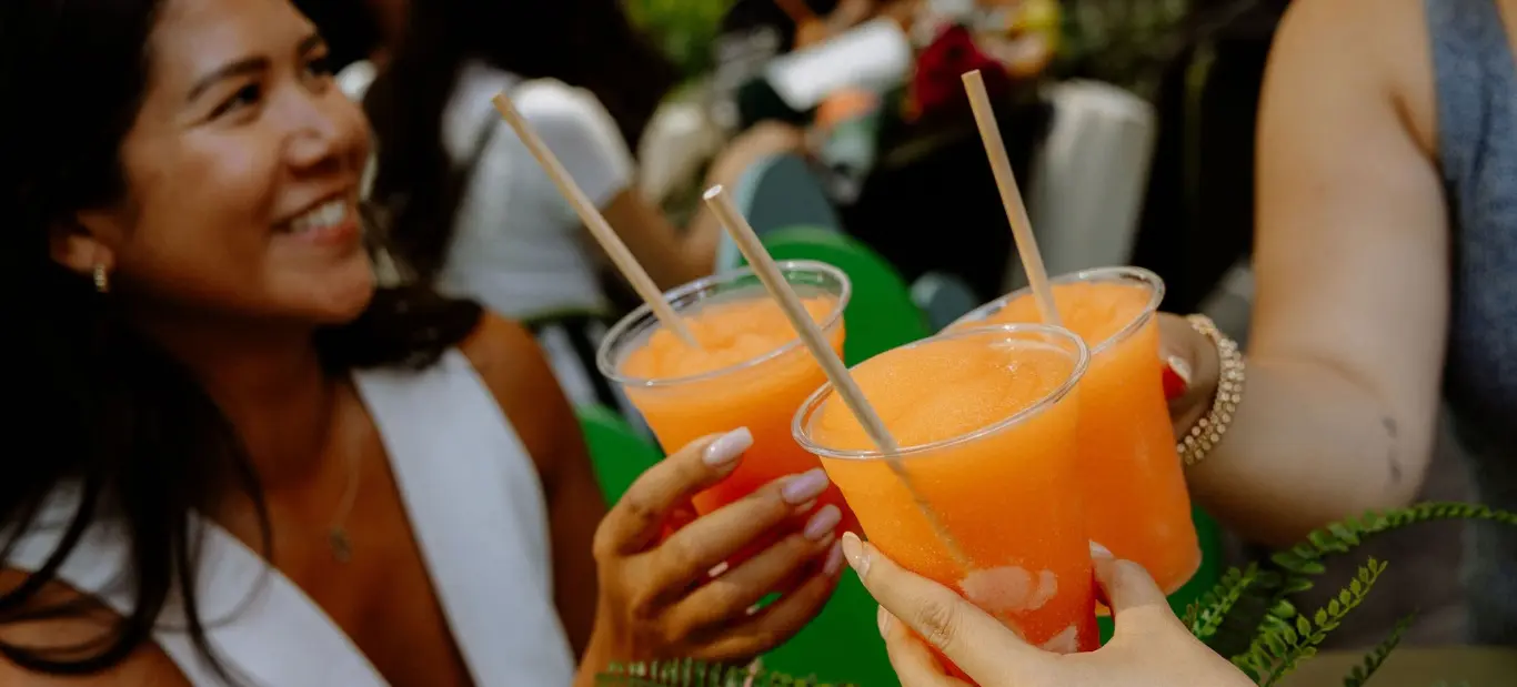 Three people holding and clinking plastic cups filled with orange slush drinks with straws.