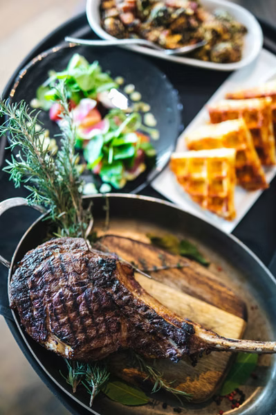 Grilled tomahawk steak garnished with rosemary in a pan, with salad and waffle slices on plates in the background.