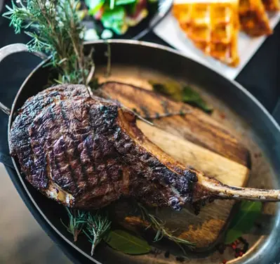 Grilled tomahawk steak garnished with rosemary in a pan, with salad and waffle slices on plates in the background.