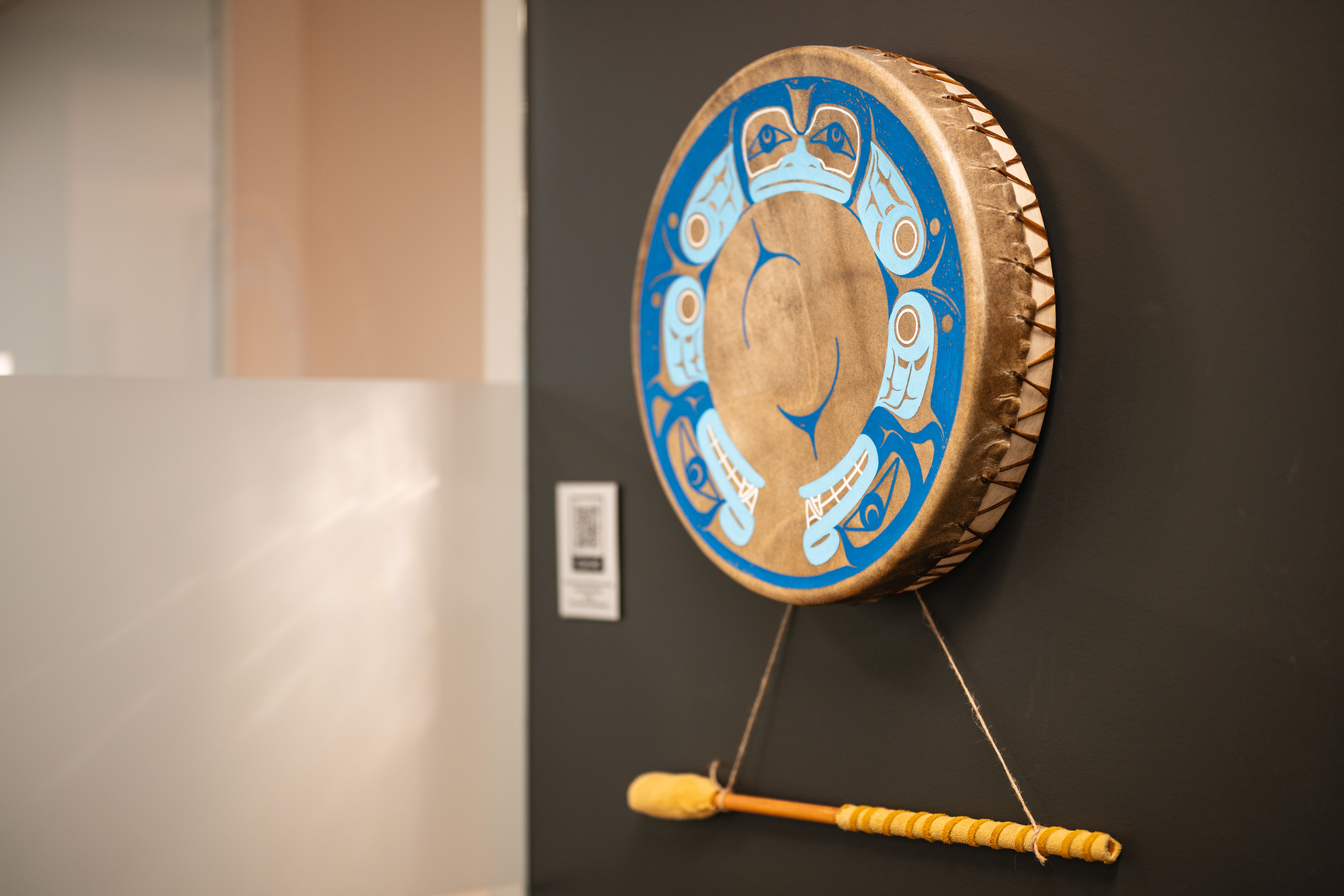 Round drum with blue and white indigenous art design hanging on a dark wall with a drumstick below it.