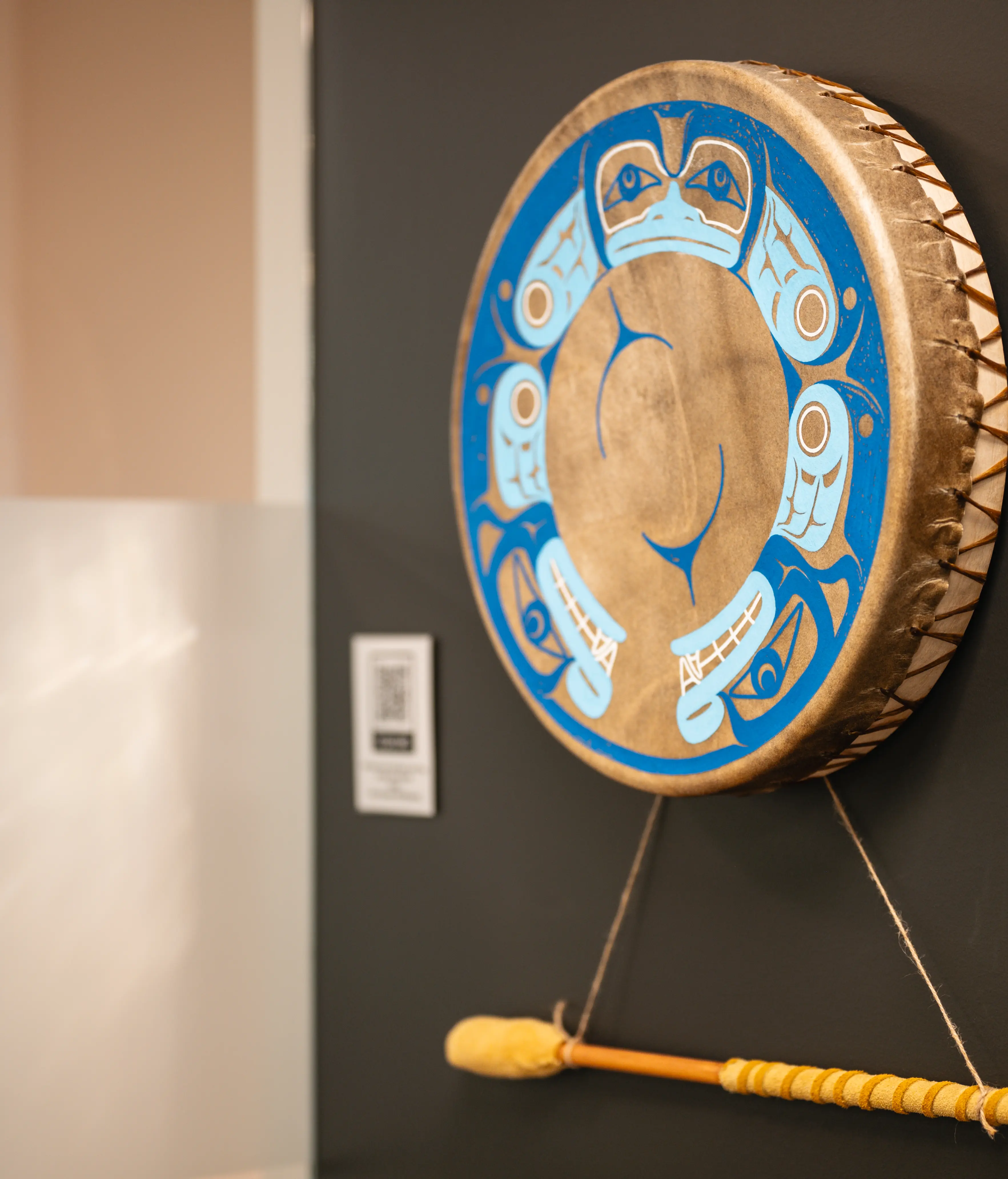 Round drum with blue and white indigenous art design hanging on a dark wall with a drumstick below it.