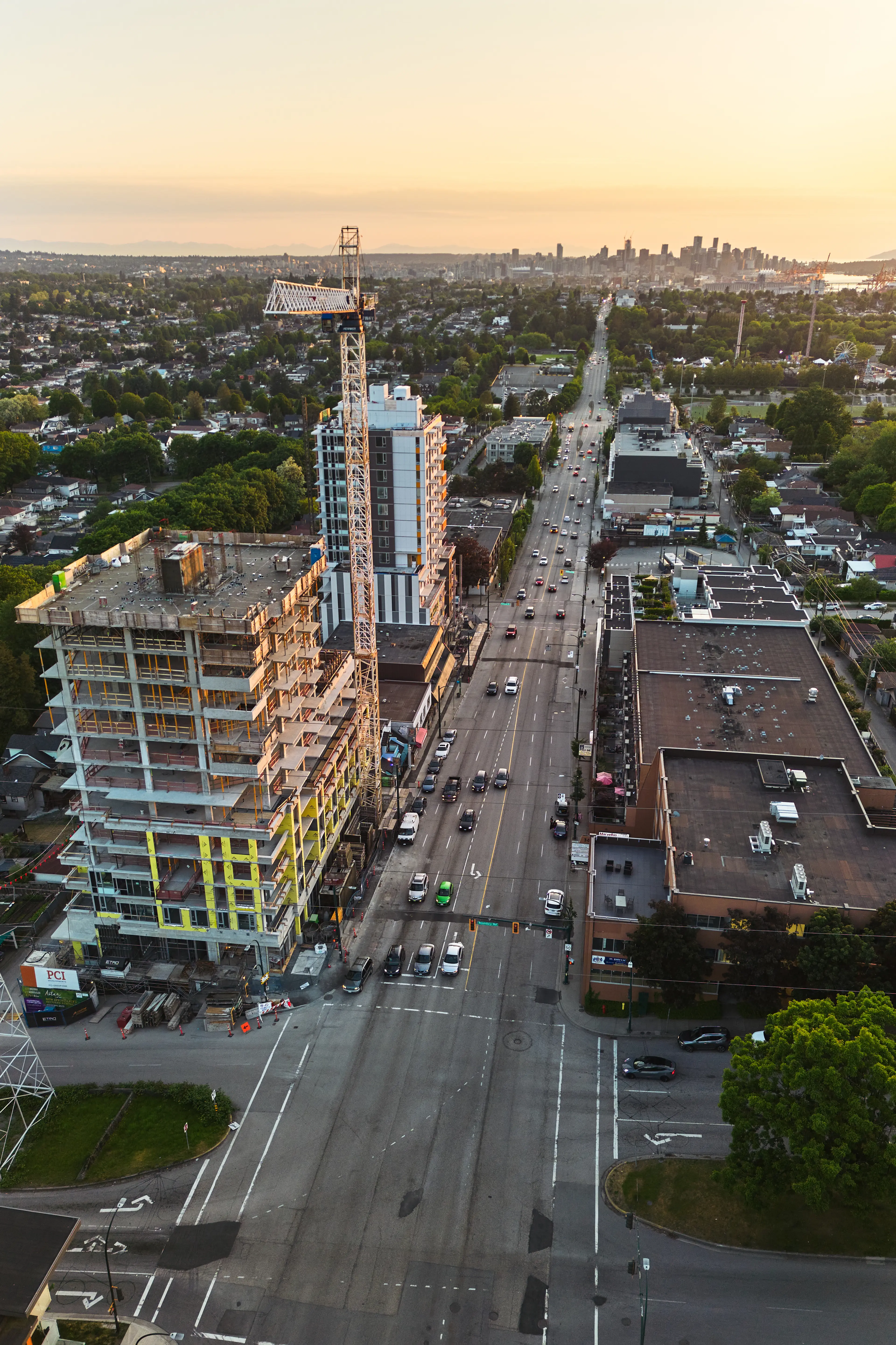Aerial view of a city street with a construction site, crane, and mid-rise buildings at sunset.