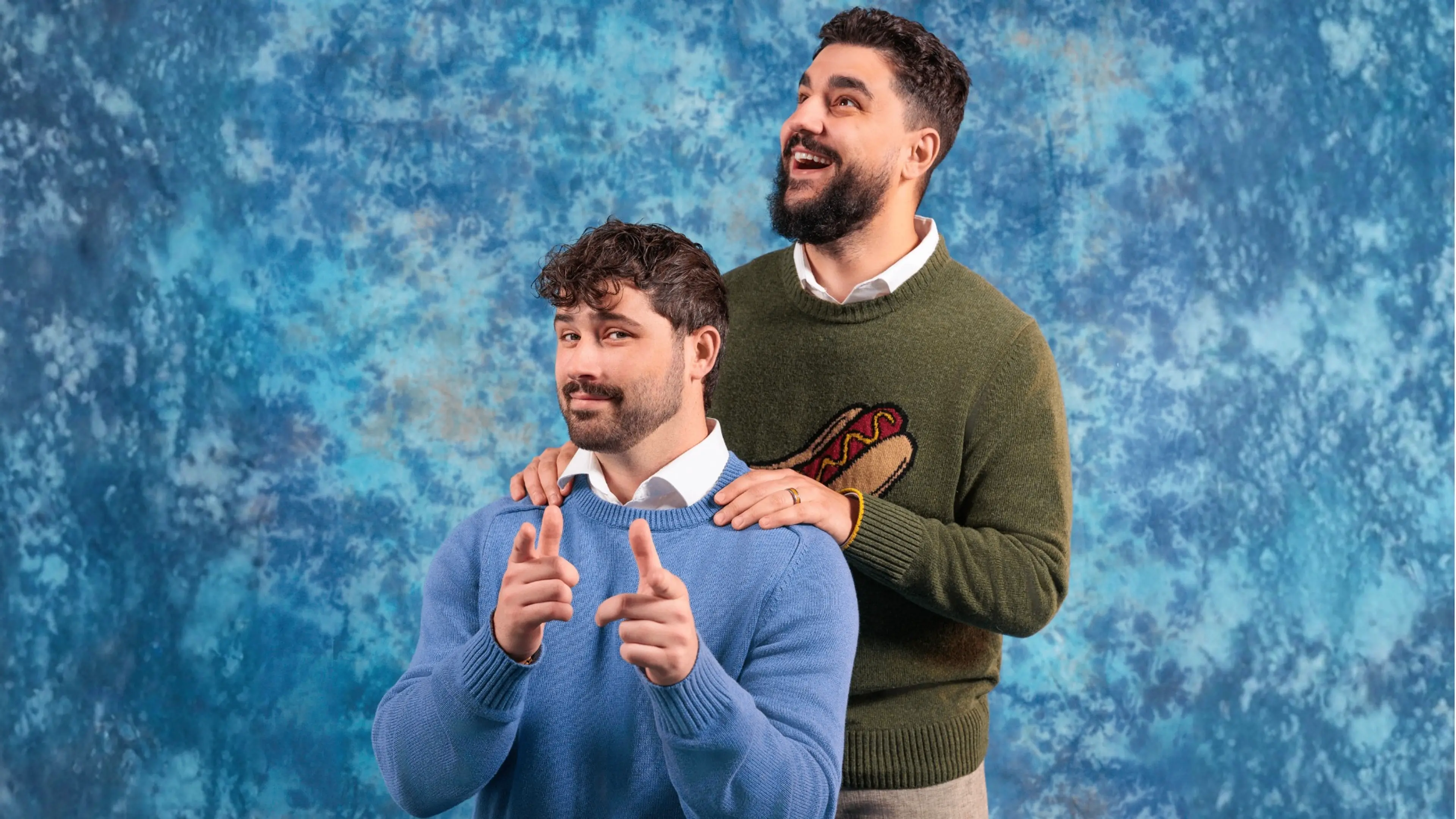 Two people with beards, one in a blue sweater pointing forward, the other in a green sweater with a hot dog design smiling and resting hands on the other's shoulders.