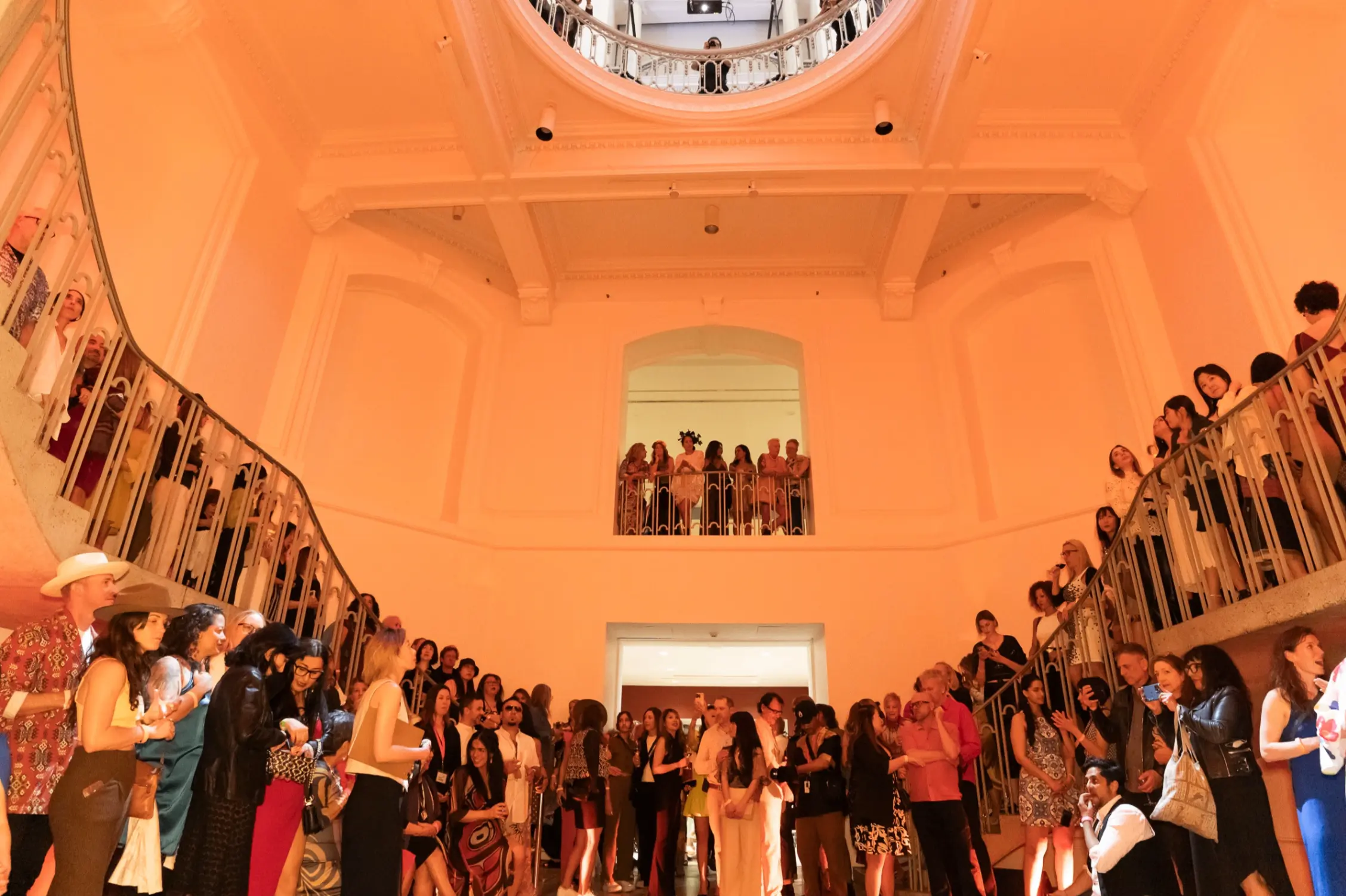 Large group of people gathered in a spacious, warmly lit room with balconies and a circular skylight above.