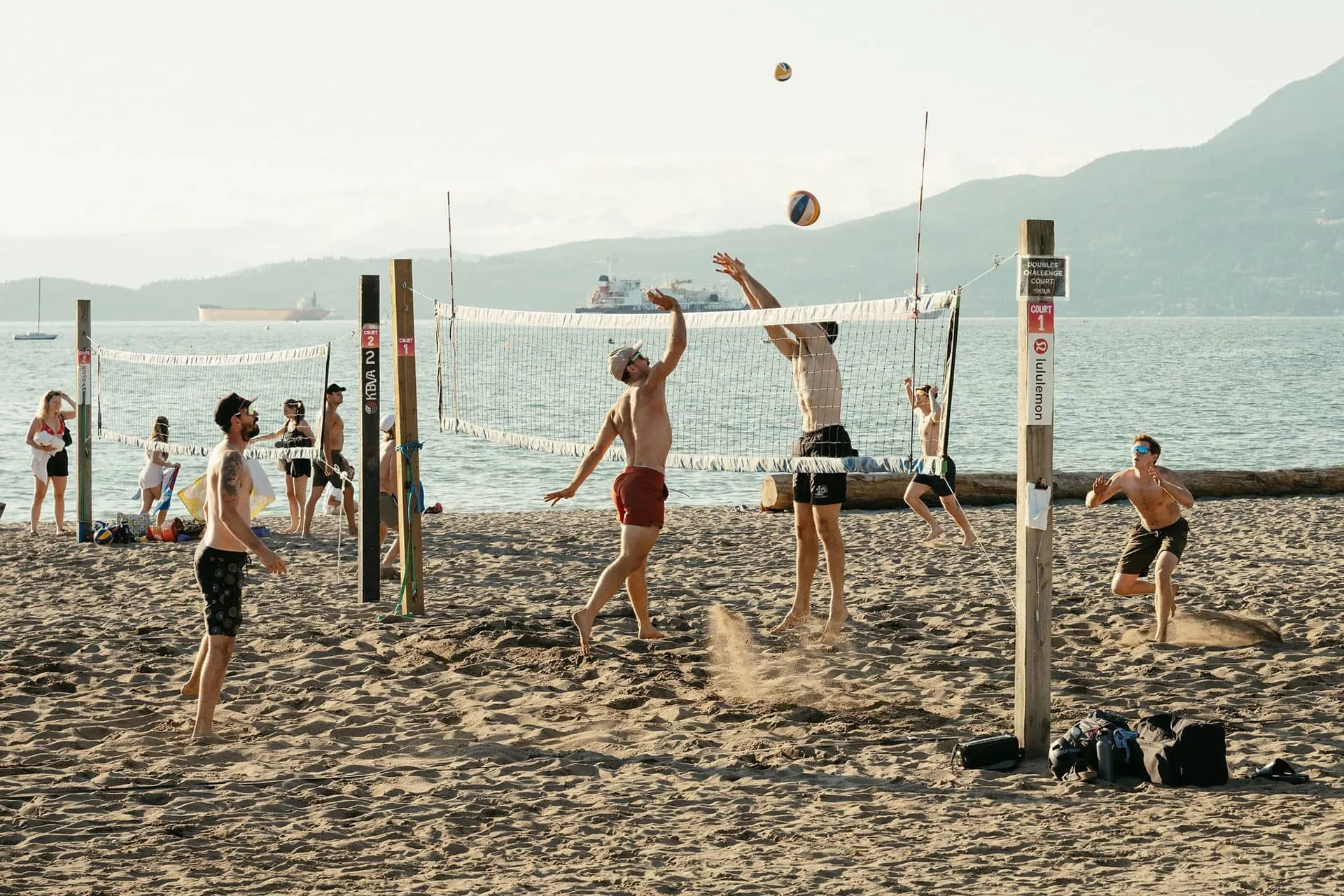 Six people playing beach volleyball on sand courts near the water with mountains and boats in the background.