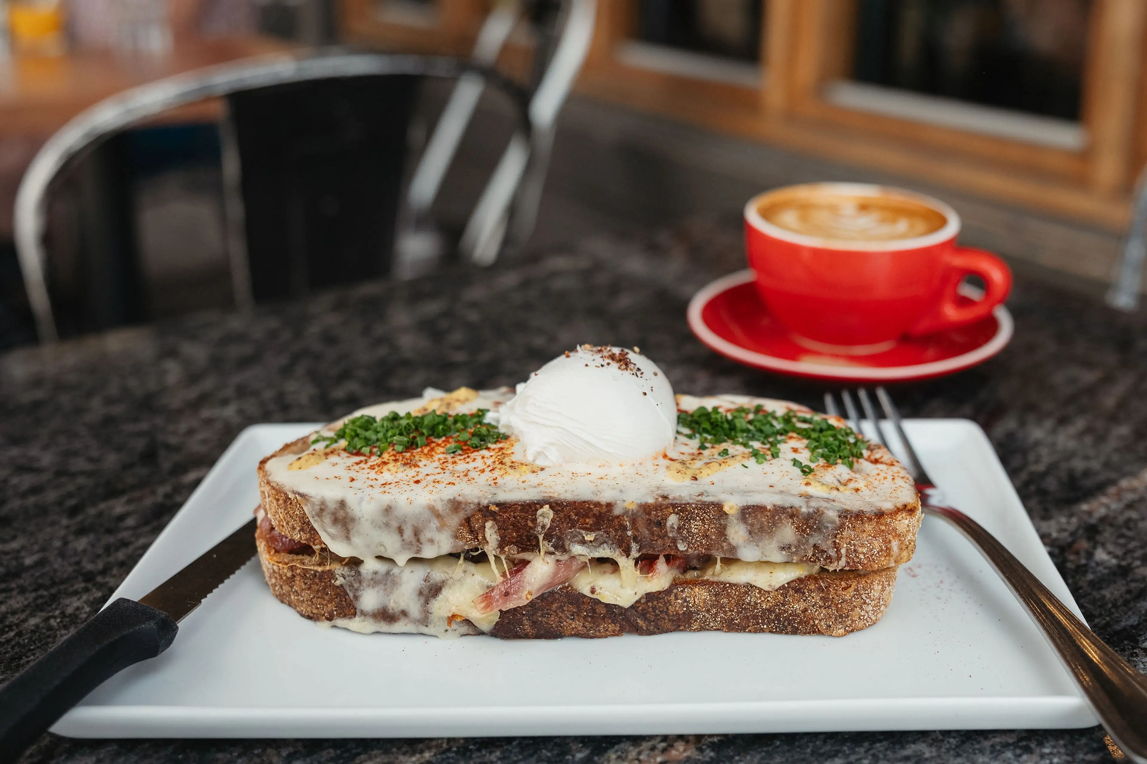 Croque madame sandwich with melted cheese, ham, herbs, and a poached egg on top, served on a white plate with a knife and fork, and a red cup of coffee in the background.
