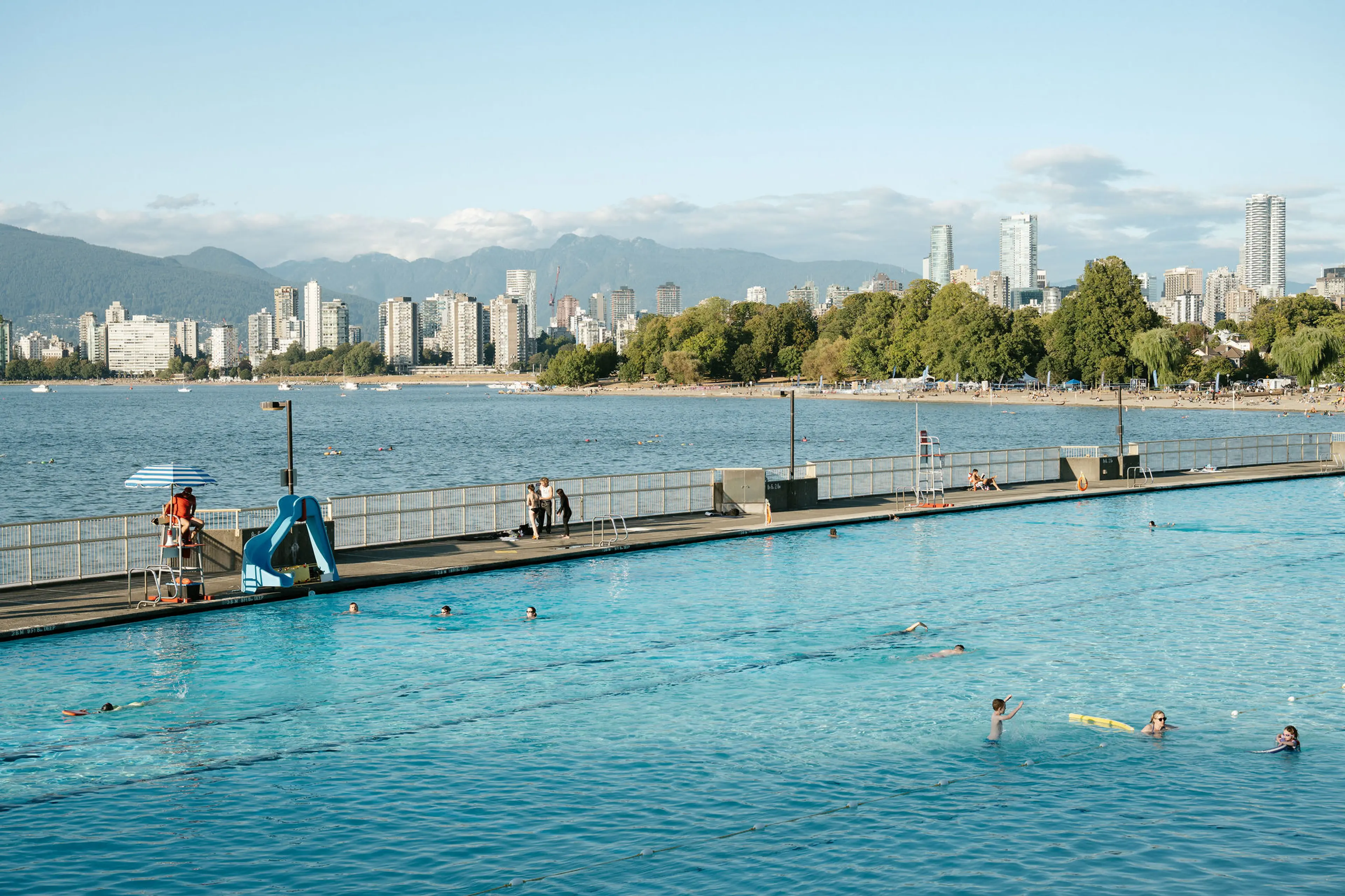 Outdoor swimming pool with people swimming and relaxing, city skyline and mountains in the background.