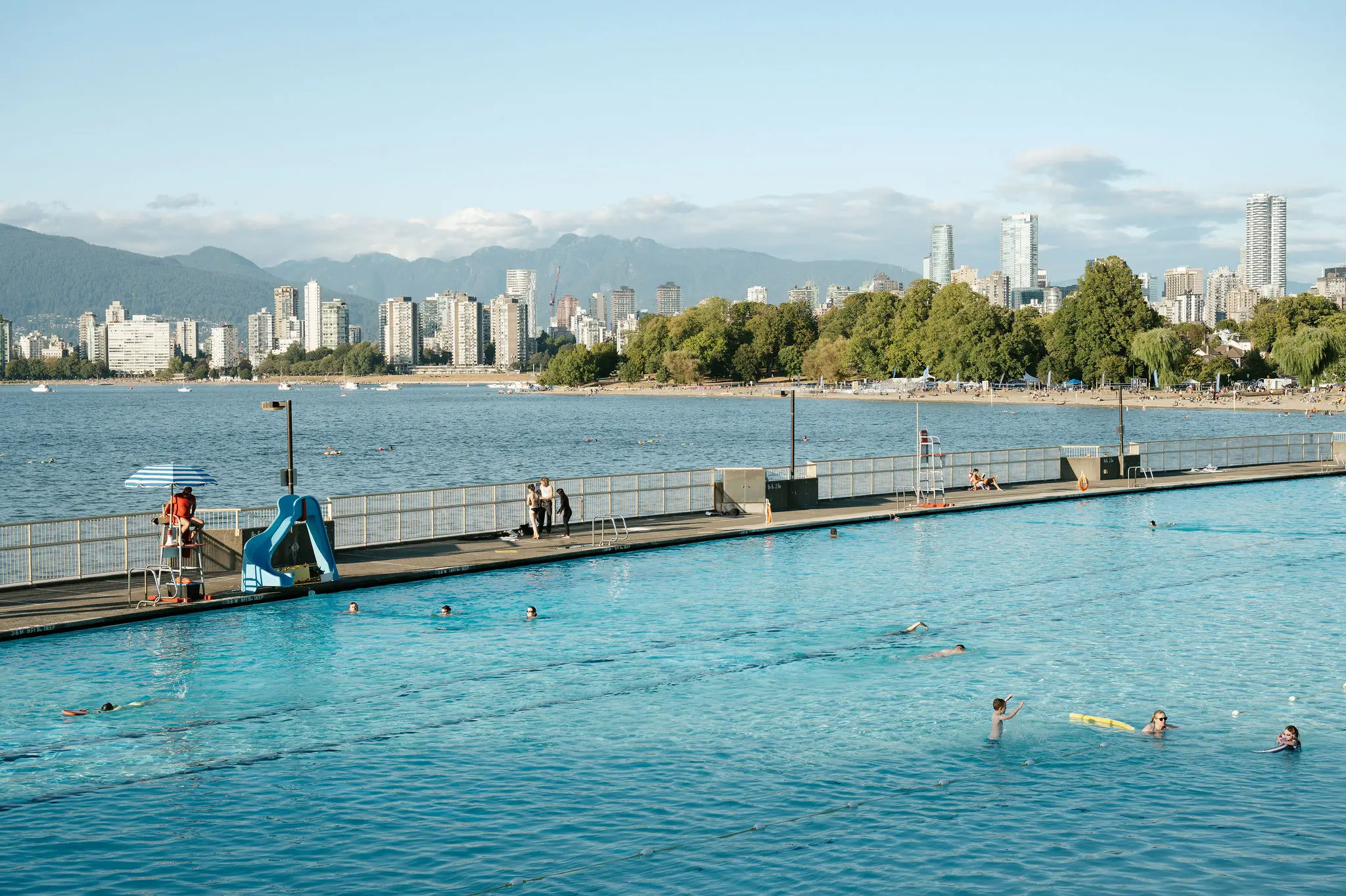 Outdoor swimming pool with people swimming and relaxing, city skyline and mountains in the background.