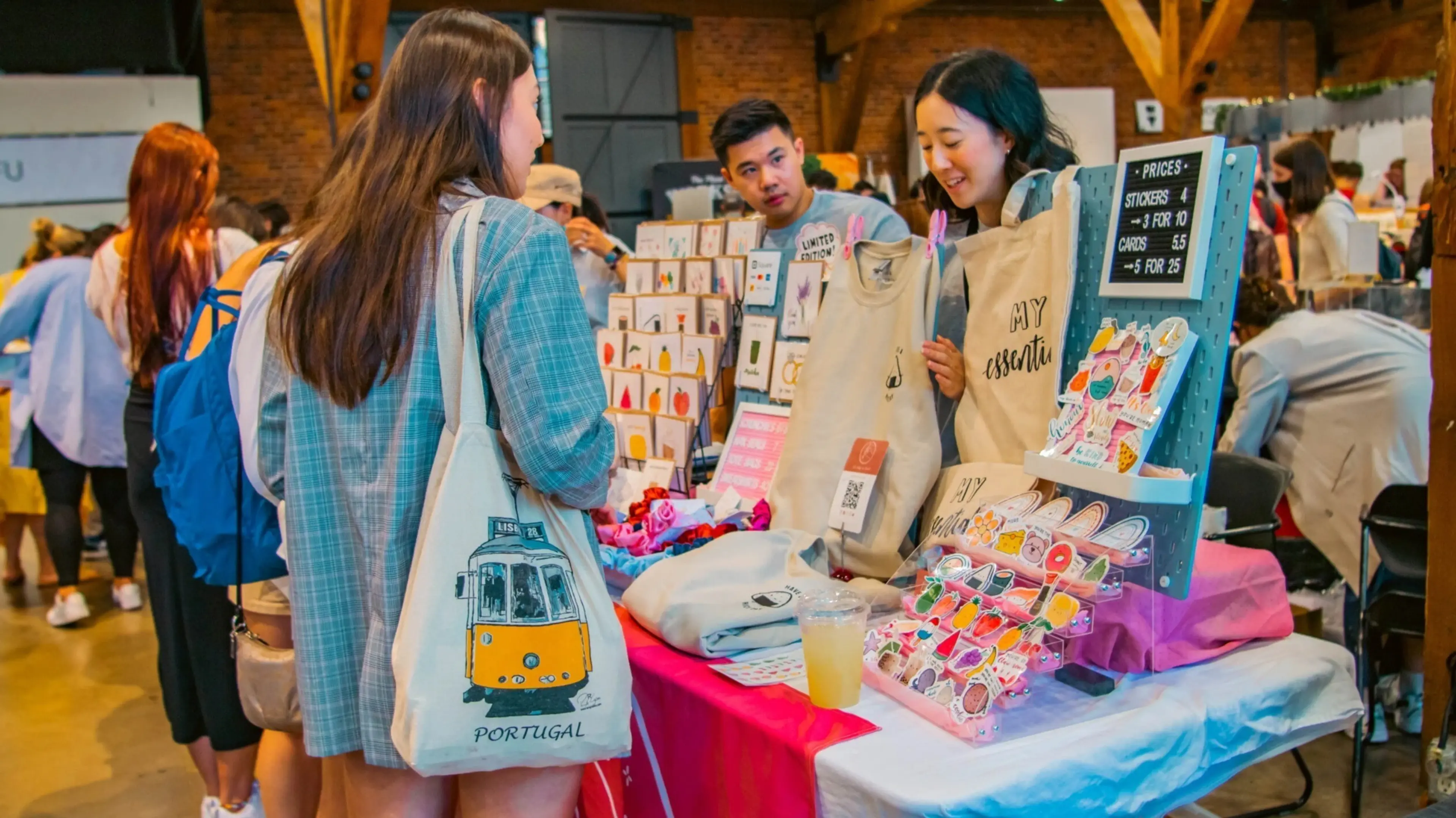 A person with a Portugal tote bag shops at a market stall selling stickers, cards, and tote bags with prices displayed.