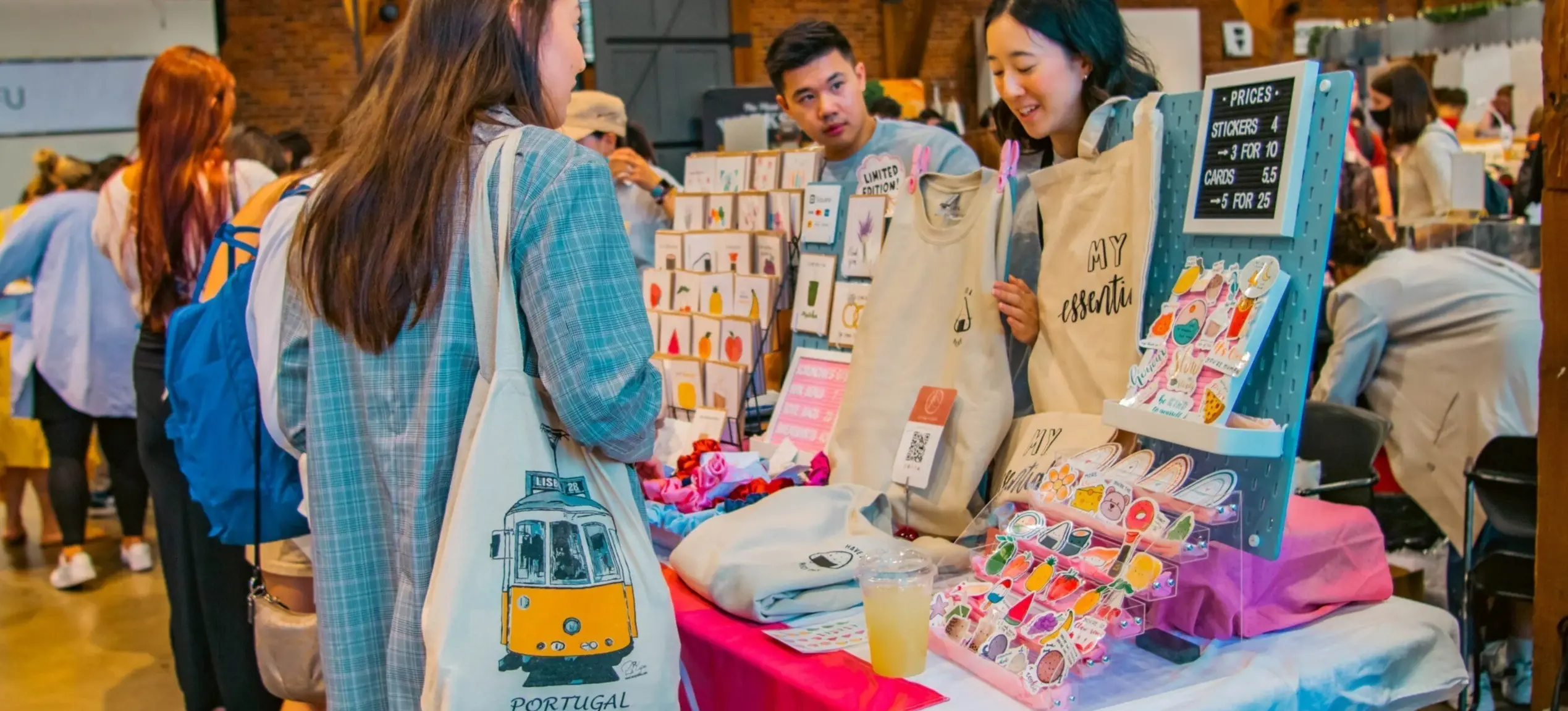 A person with a Portugal tote bag shops at a market stall selling stickers, cards, and tote bags with prices displayed.