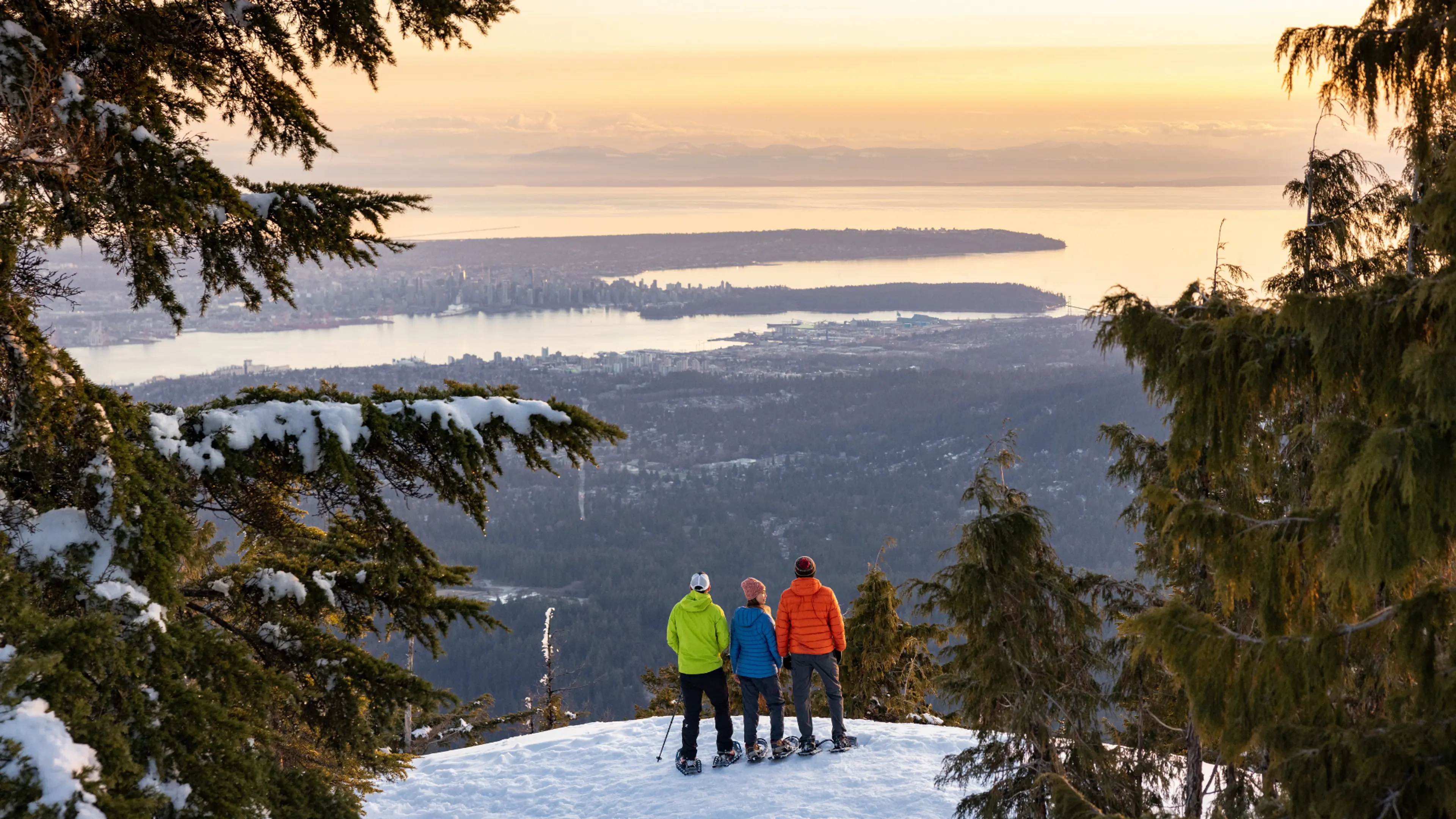 Three people in winter clothing standing on snow with snowshoes, overlooking a city and water at sunset.
