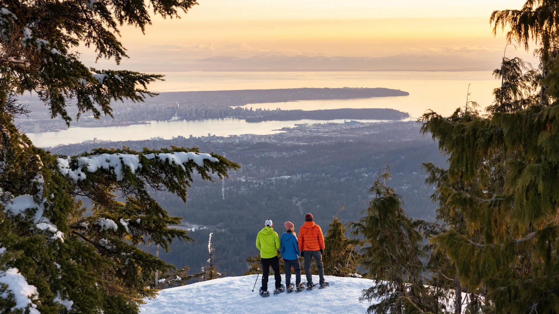 Three people in winter clothing standing on snow with snowshoes, overlooking a city and water at sunset.