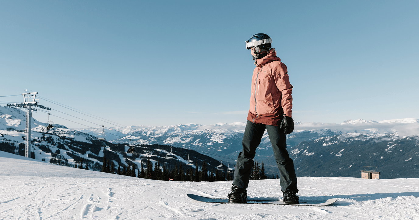 A person wearing a helmet and orange jacket snowboarding on a snowy mountain with ski lifts and snow-covered peaks in the background.