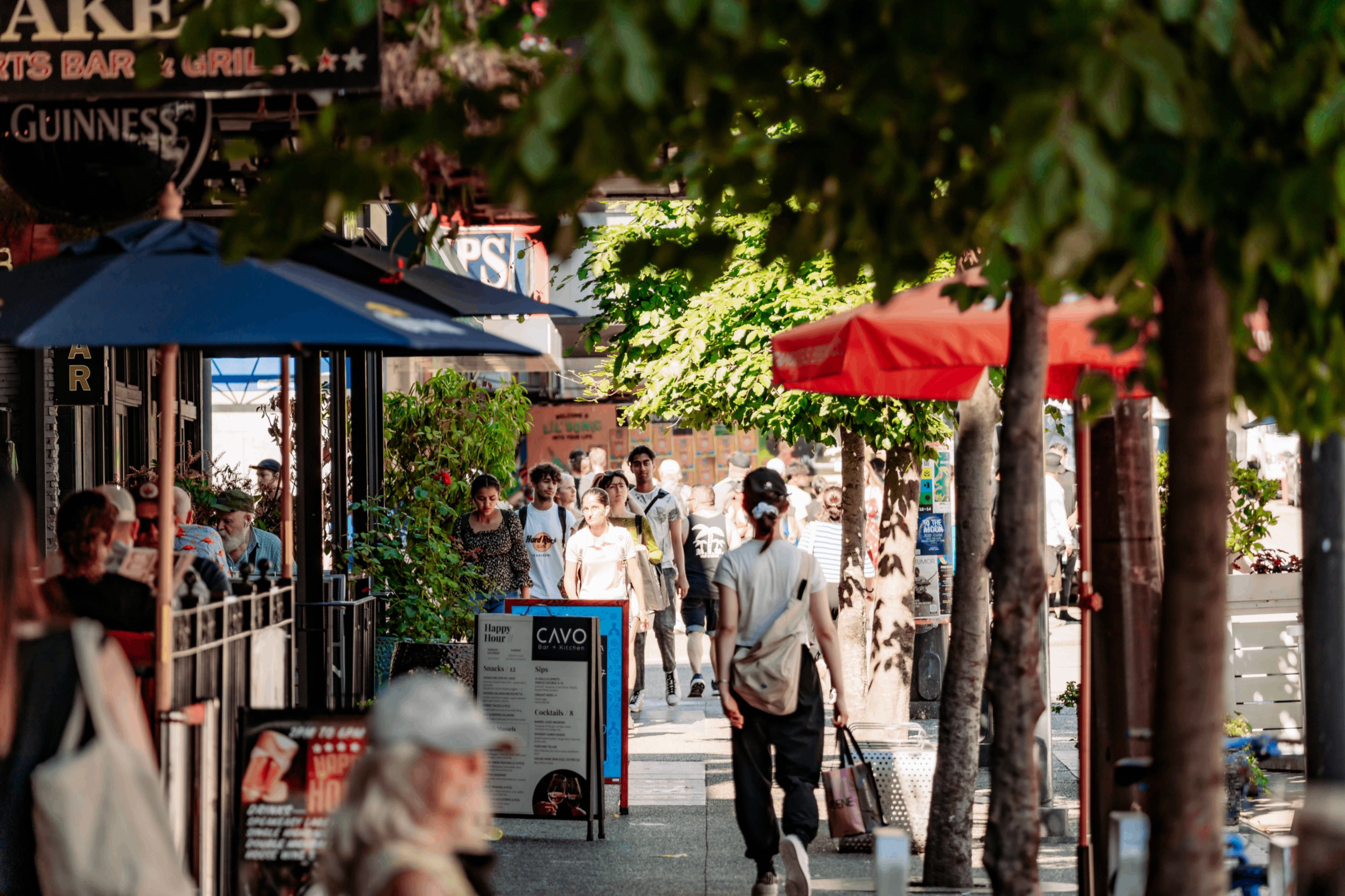 People walking on a tree-lined city sidewalk with outdoor seating and umbrellas at cafes and restaurants.