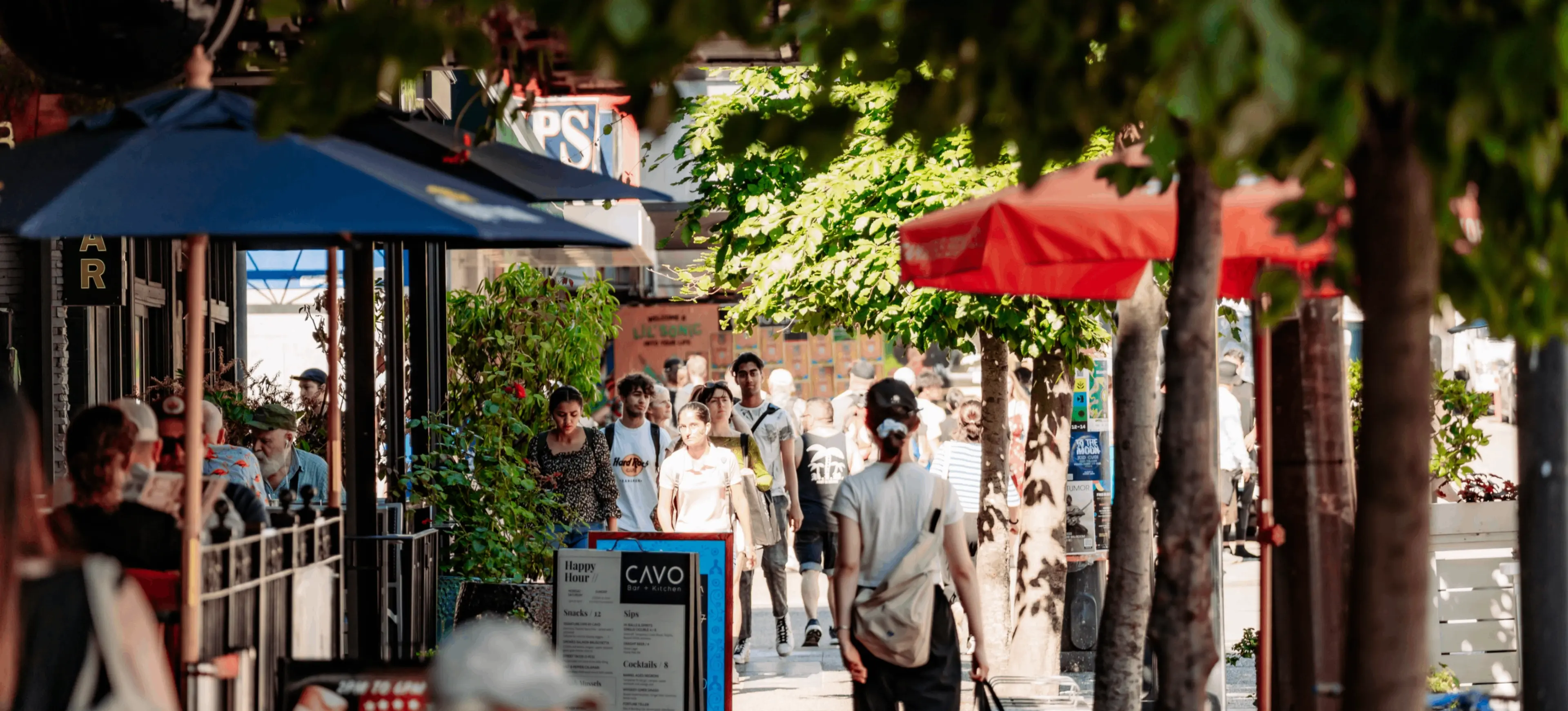 People walking on a tree-lined city sidewalk with outdoor seating and umbrellas at cafes and restaurants.